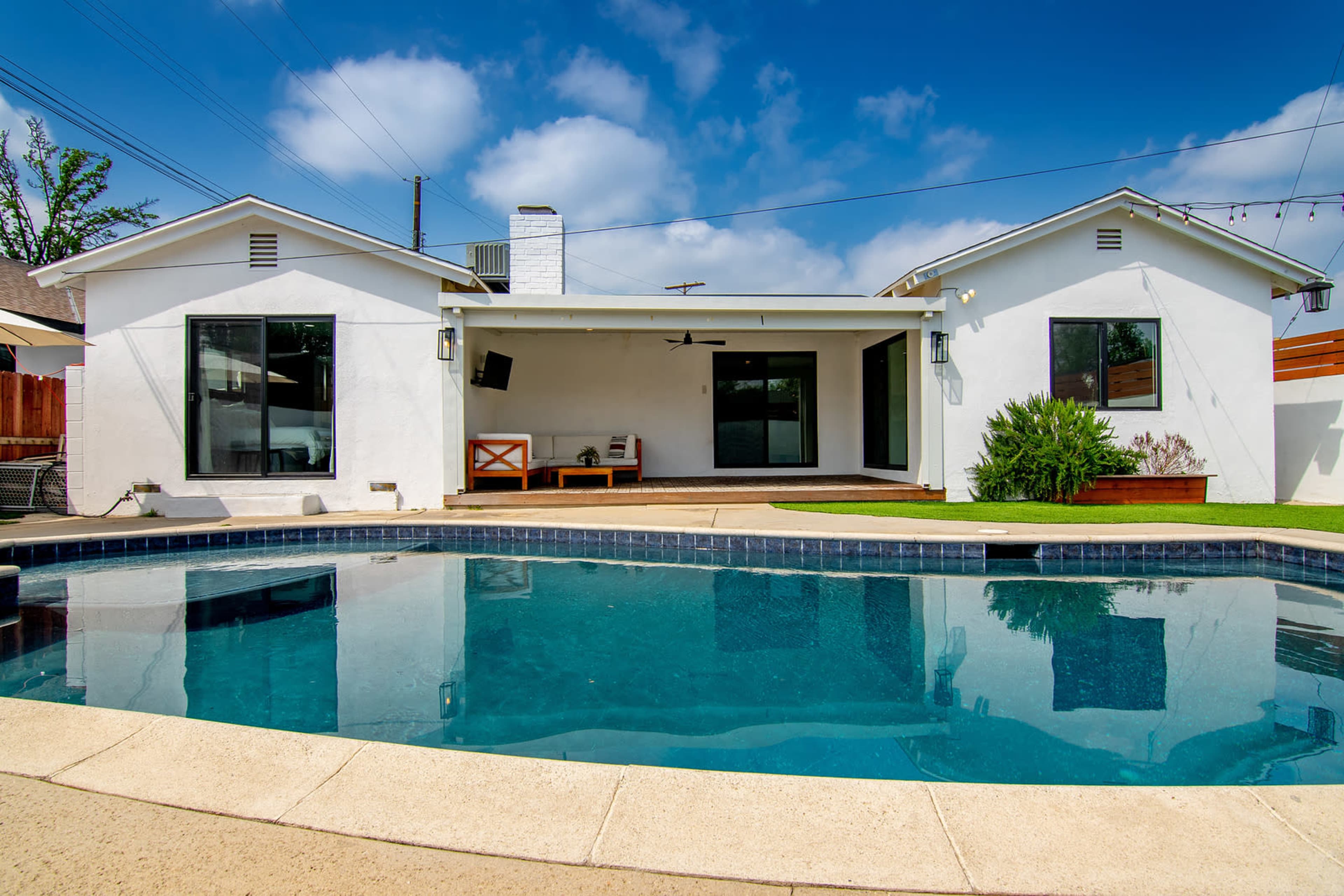 A white house with large windows and sliding doors, overlooking a swimming pool surrounded by a patio and grass lawn under a blue sky.