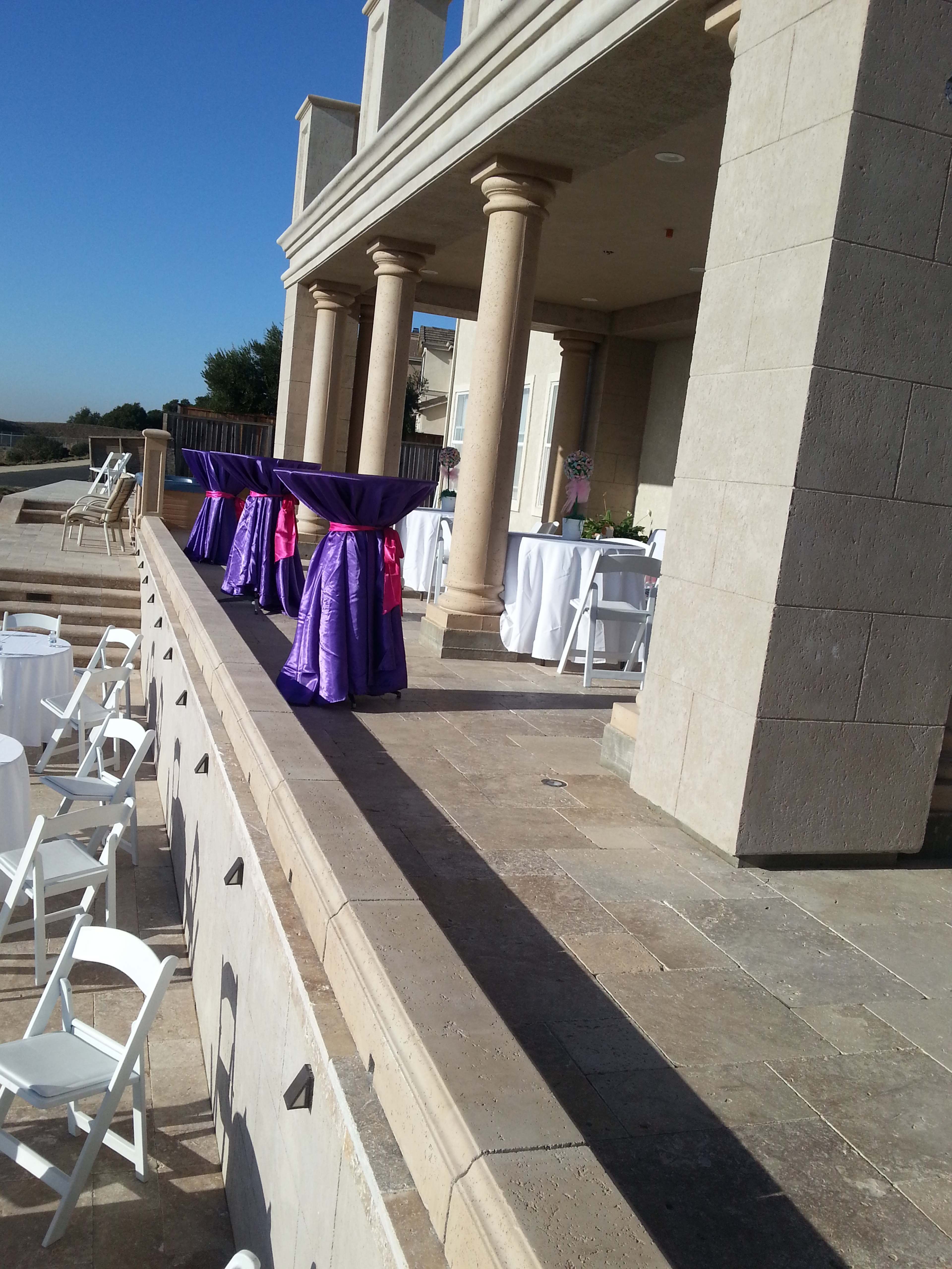 The image shows a patio area with columns, featuring tables draped in purple and pink tablecloths, set up for an event.