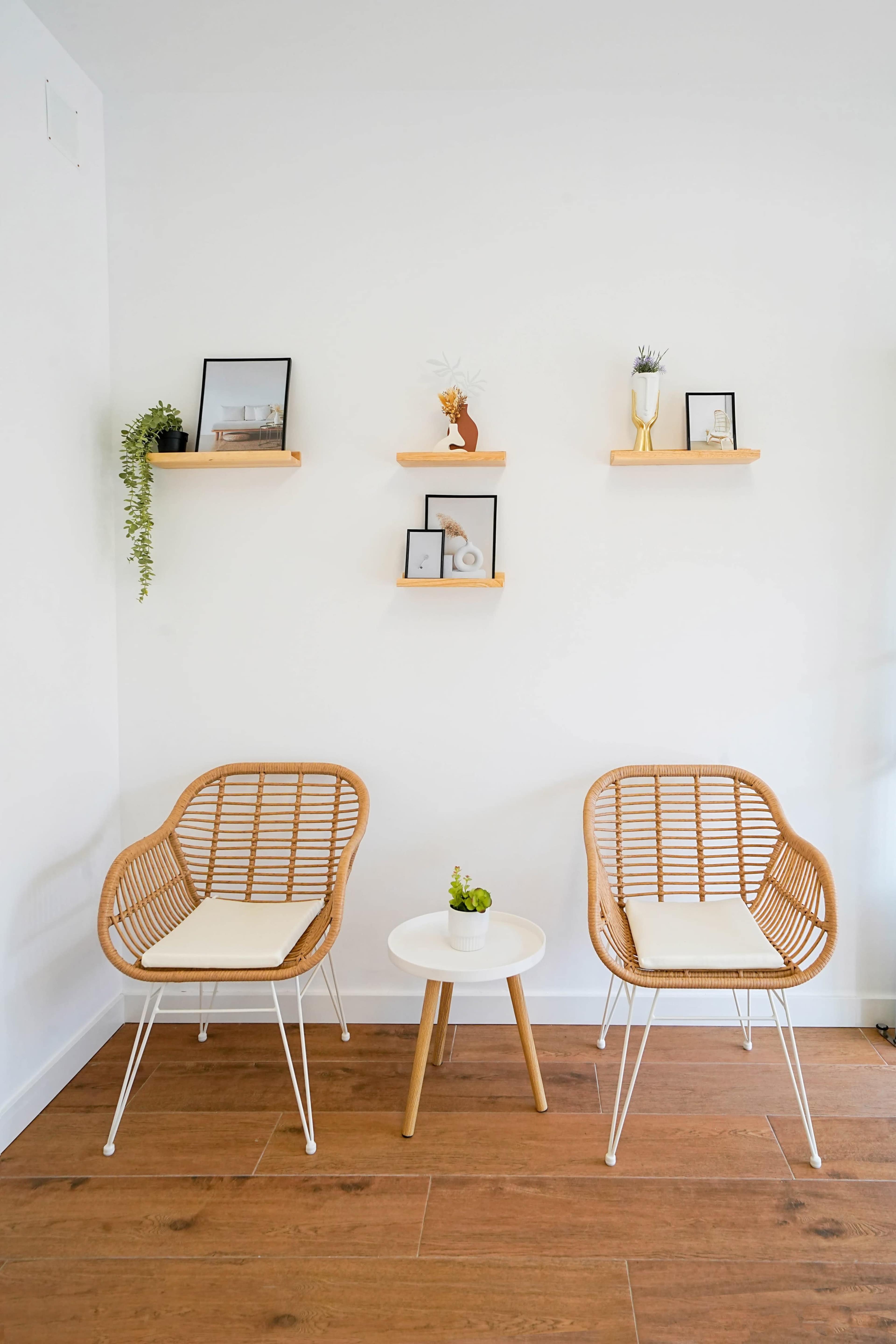 The image shows a minimalistic seating area with two woven chairs, a small round table, and a few decorative shelves on the wall holding plants and framed pictures.