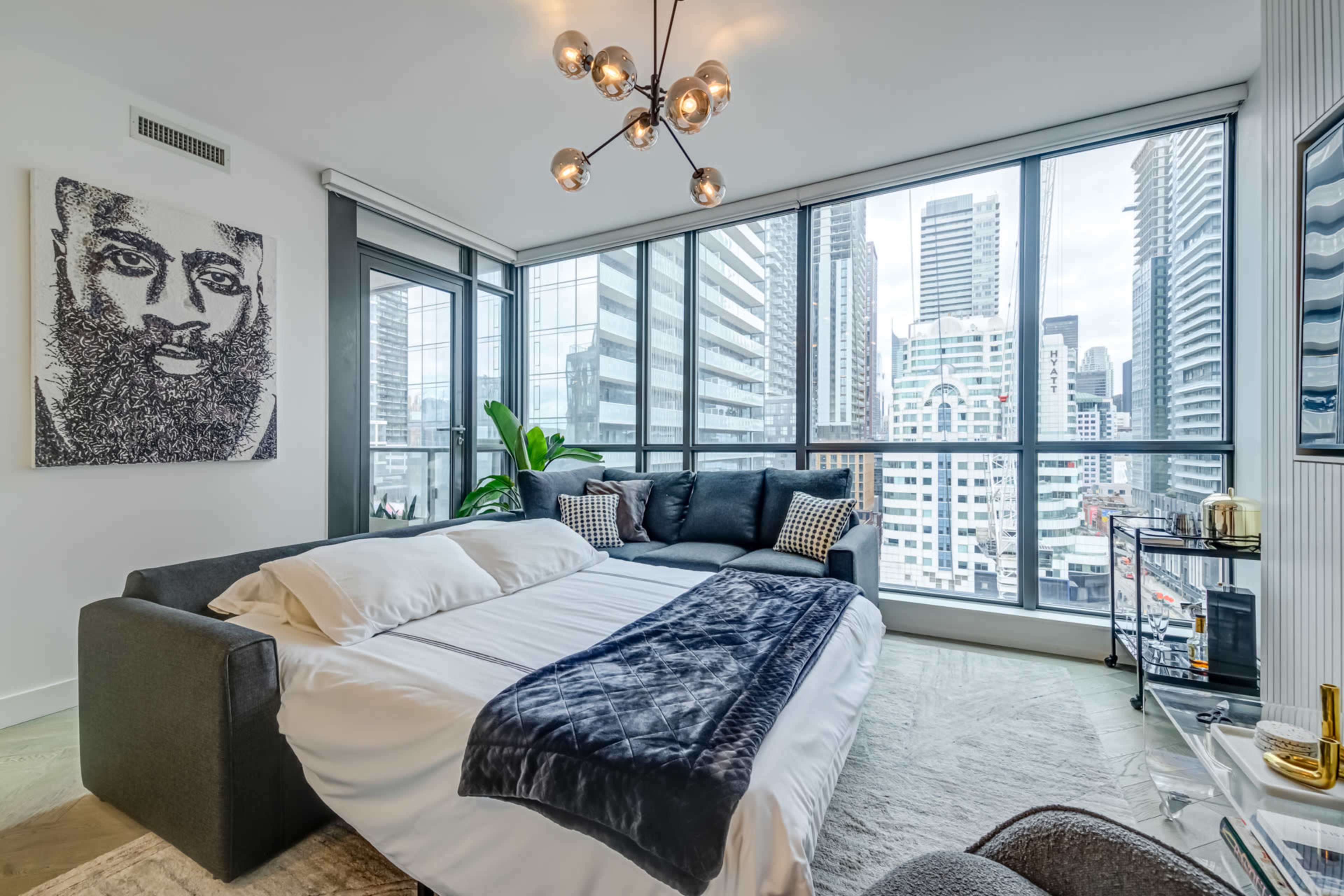 The image shows a modern living area with a sofa bed, large windows revealing a city skyline, and a wall art piece of a person's face.