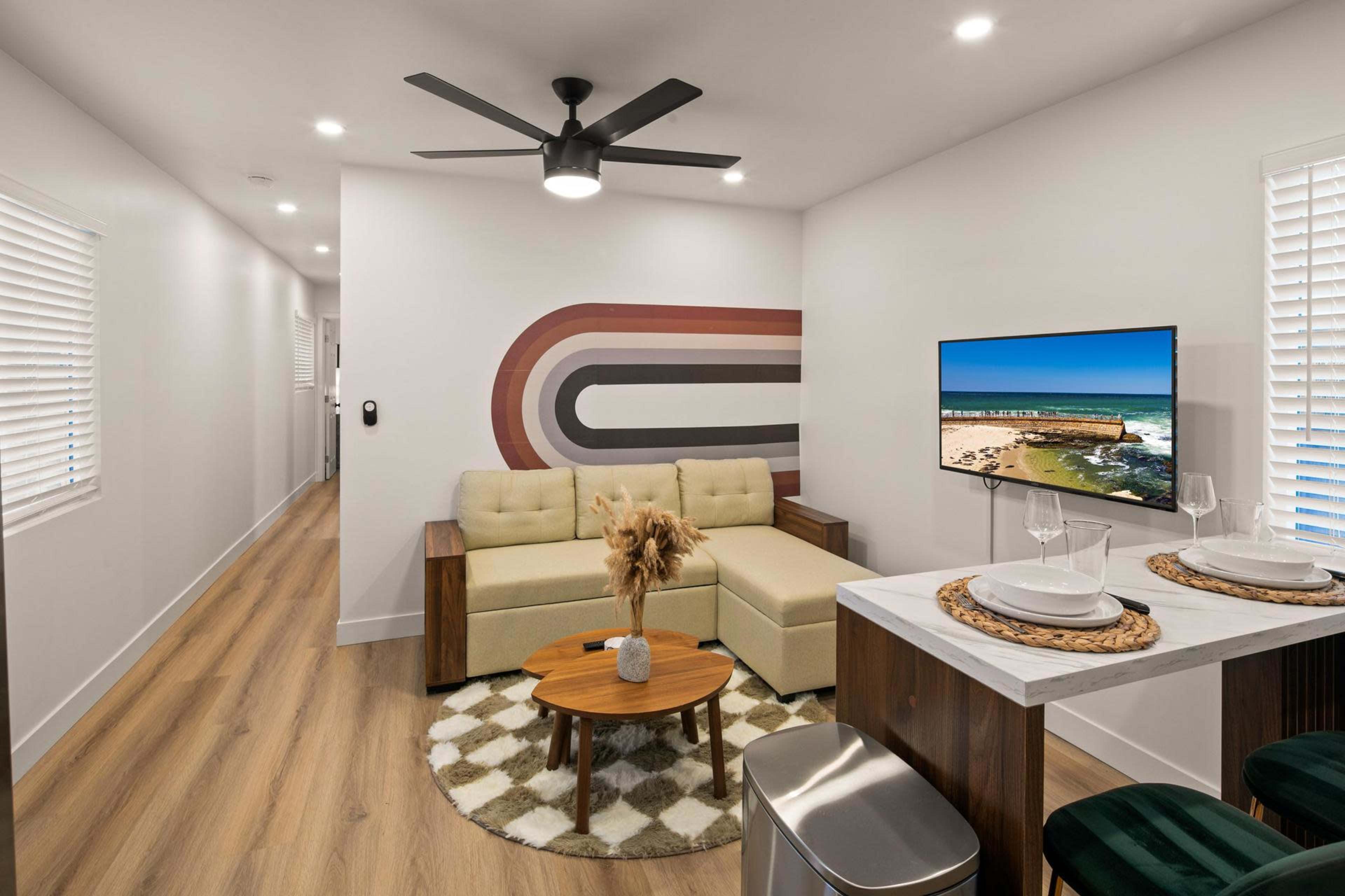 The image shows a modern living space featuring a light-colored sectional sofa, a round wooden coffee table, a wall-mounted television displaying a landscape, and a dining area with two plates set on a marble top.