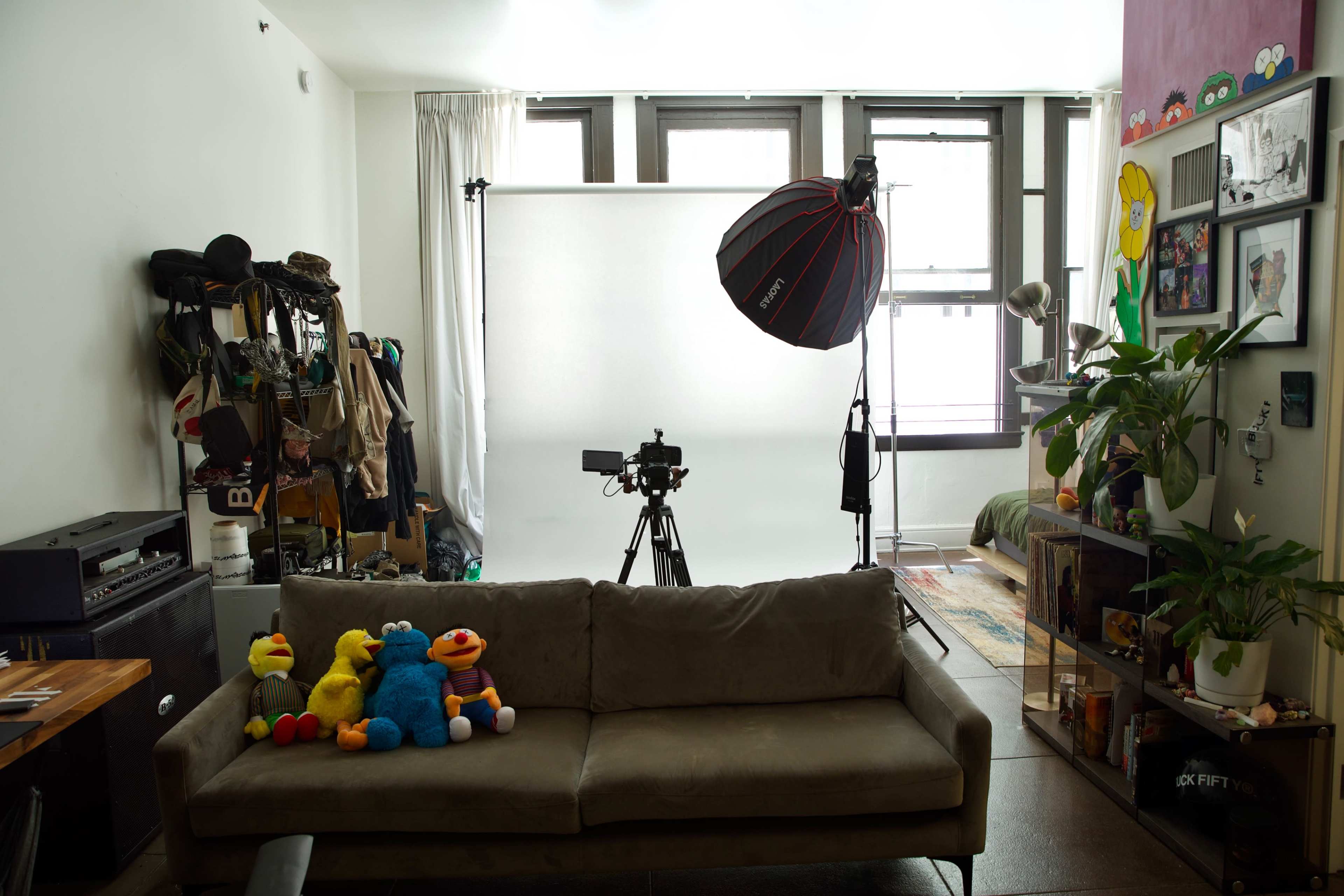 A living space featuring a couch with colorful stuffed toys, a camera setup facing a white backdrop, and various personal items arranged around the room.