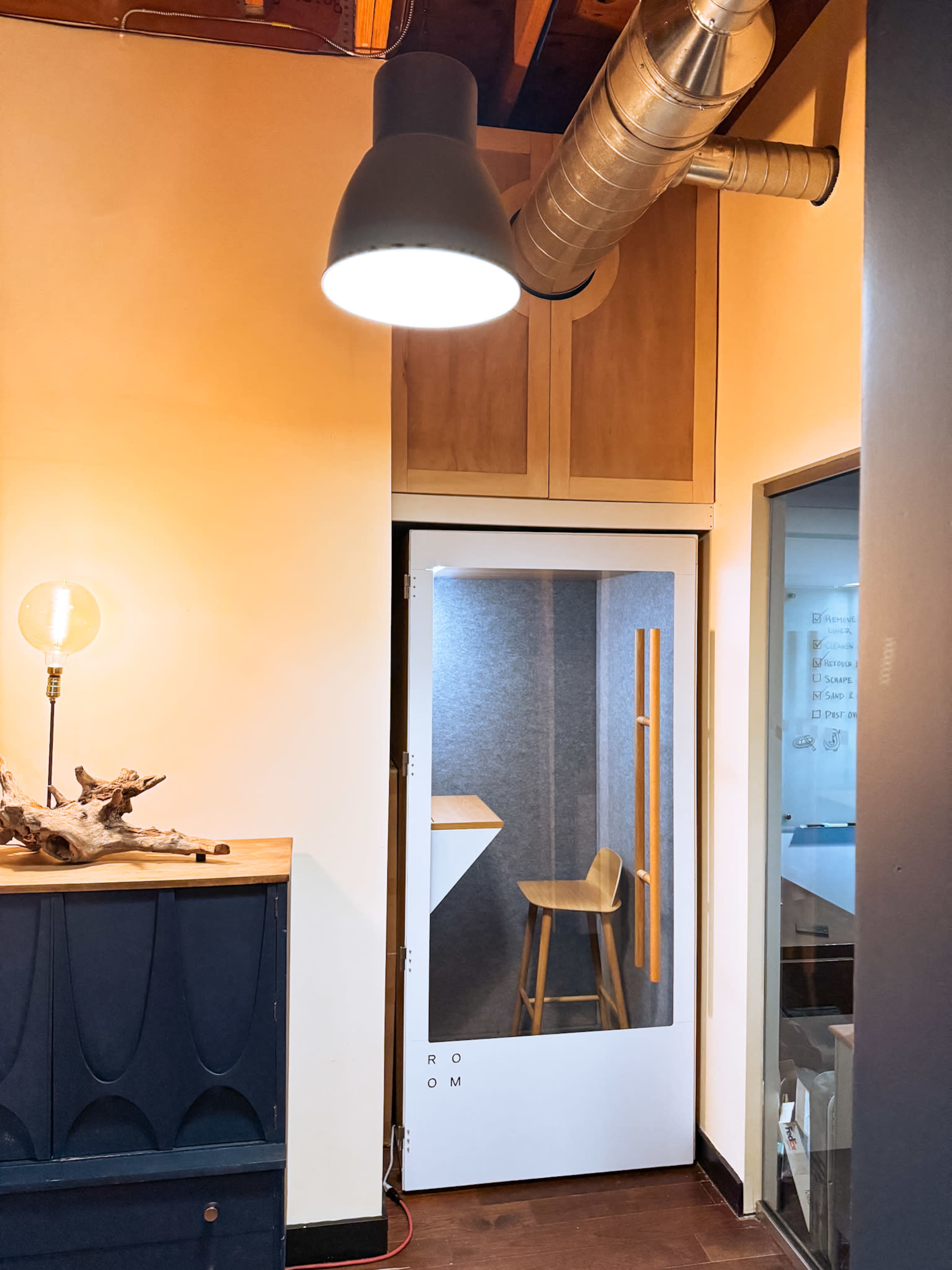 A modern office hallway features a door with a glass panel and a tall chair visible inside, along with wooden accents and industrial lighting.