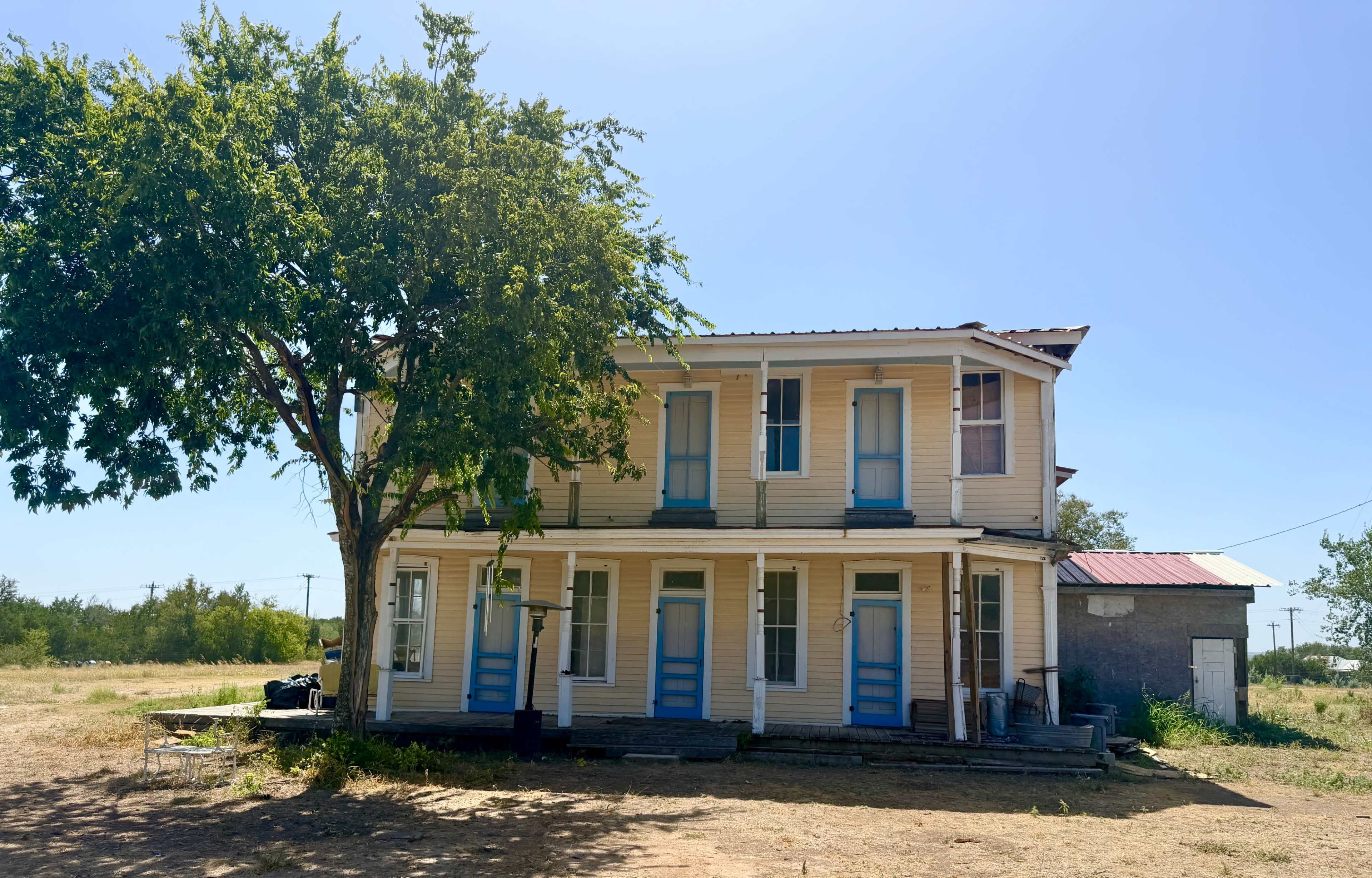 A two-story house with a light-colored exterior and blue doors stands next to a large tree in an open area.