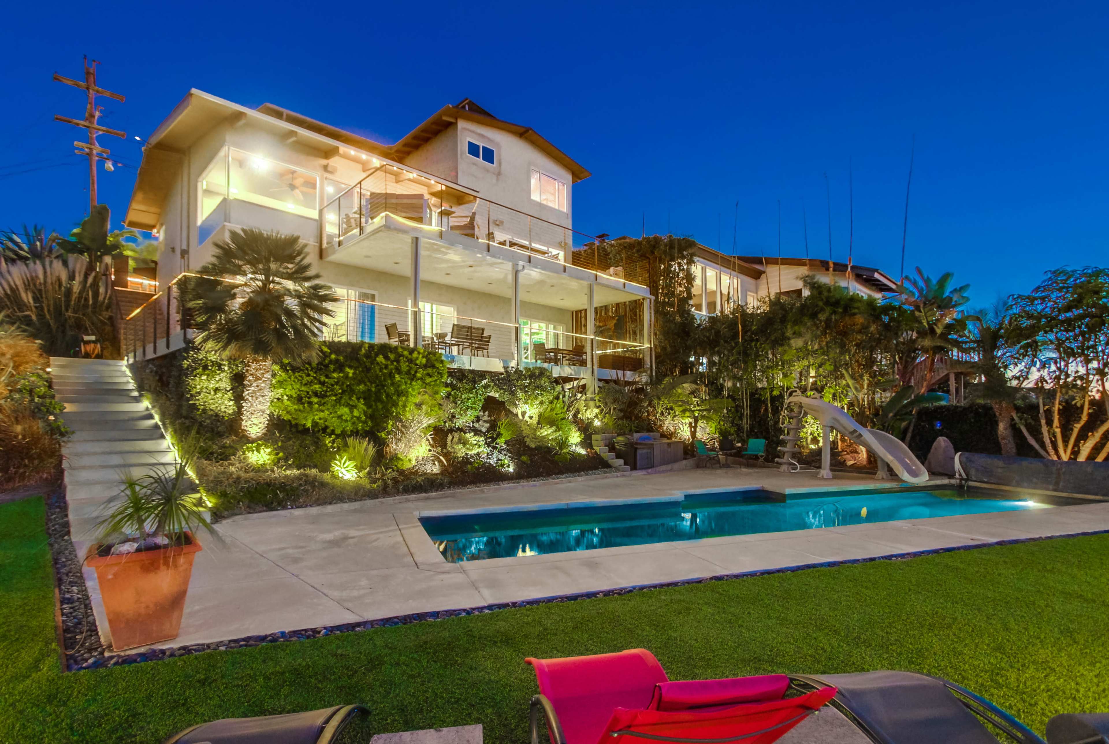 A two-story house with multiple balconies overlooks a swimming pool surrounded by landscaped greenery in the evening.