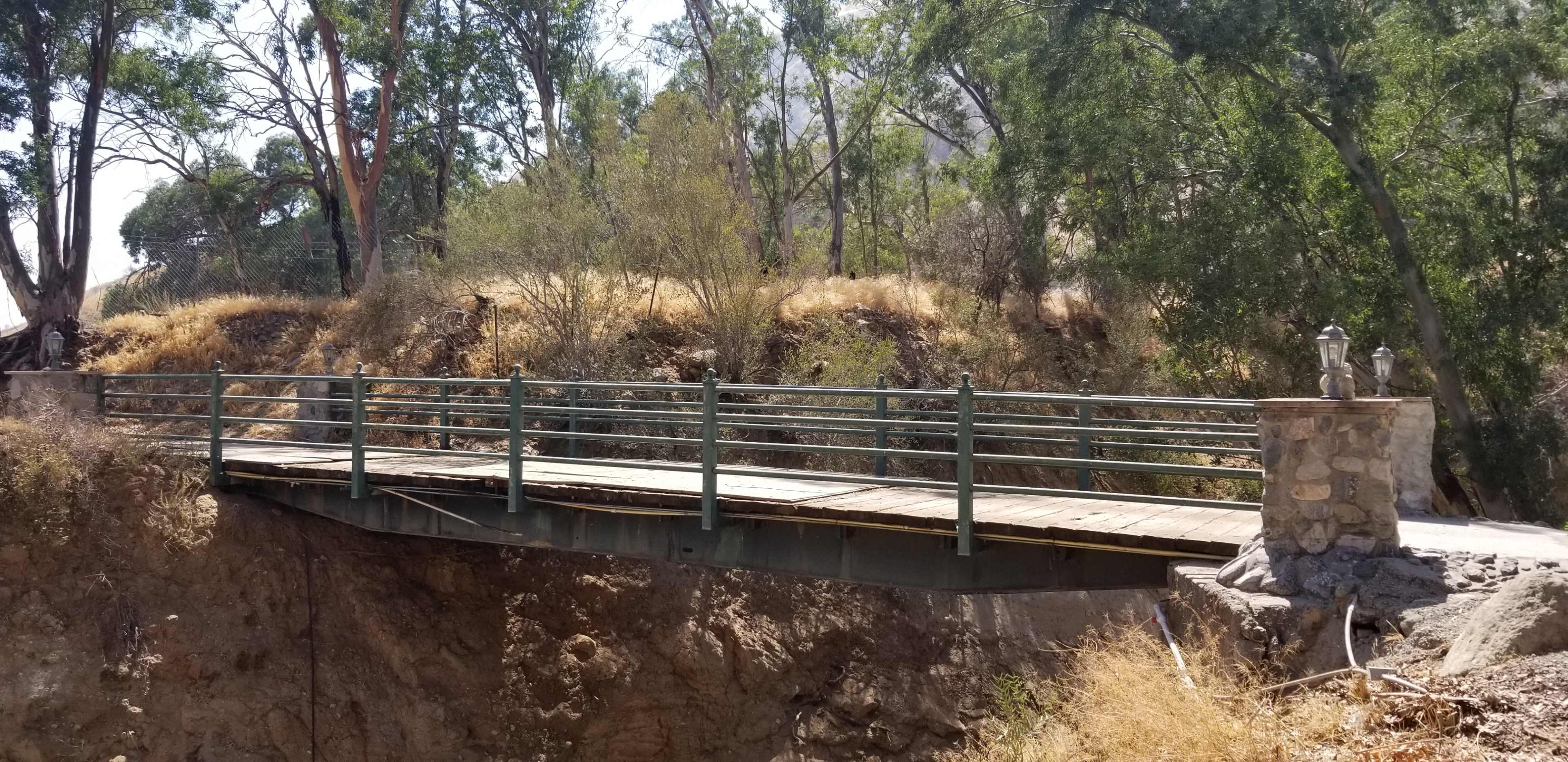 A metal bridge with wooden planks spans a dry ravine, supported by stone pillars and surrounded by trees and sparse vegetation.