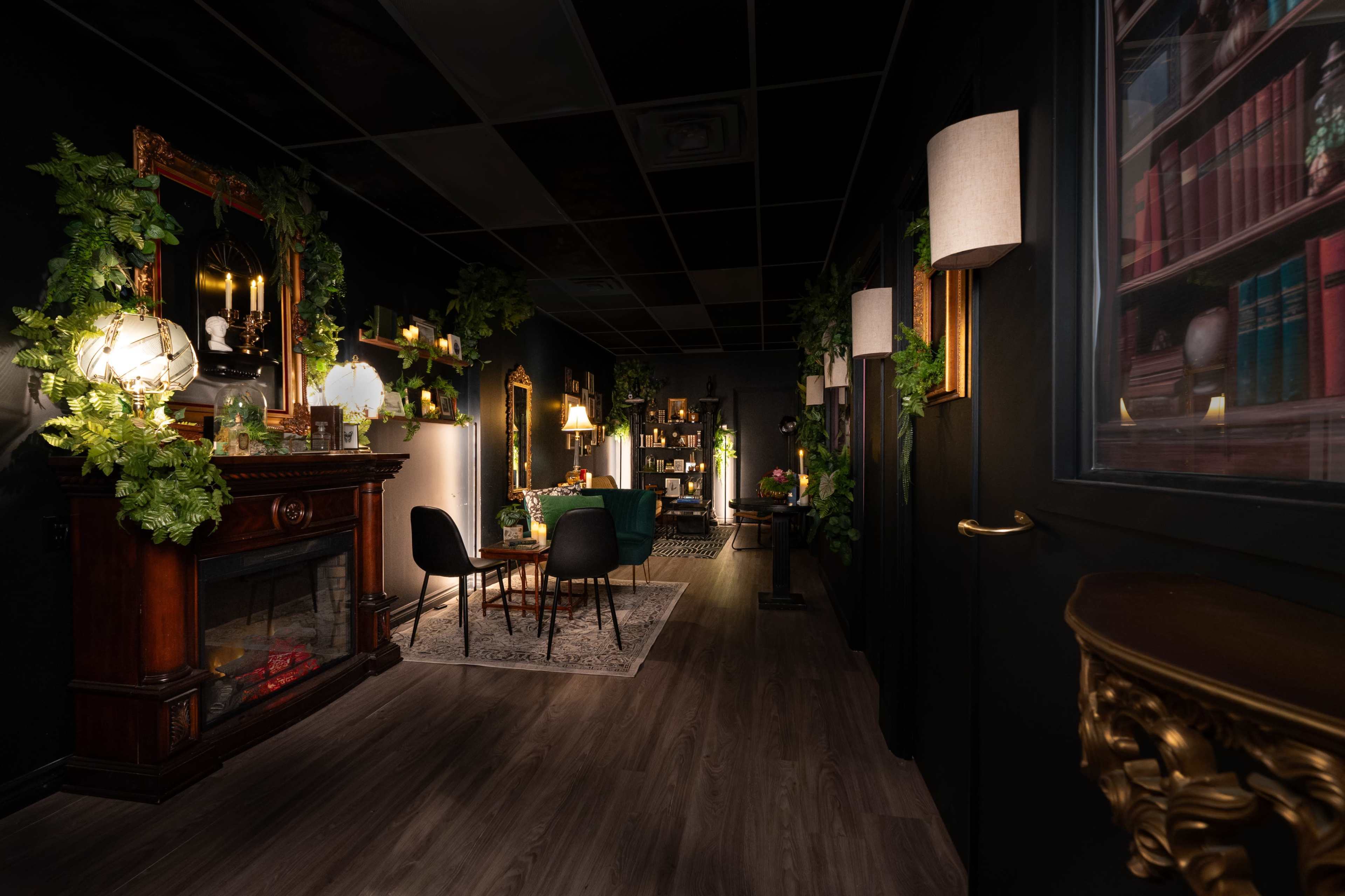 A dimly lit room with dark walls, featuring a fireplace, mirrors, various plants, and elegant furniture arranged in a cozy setting.