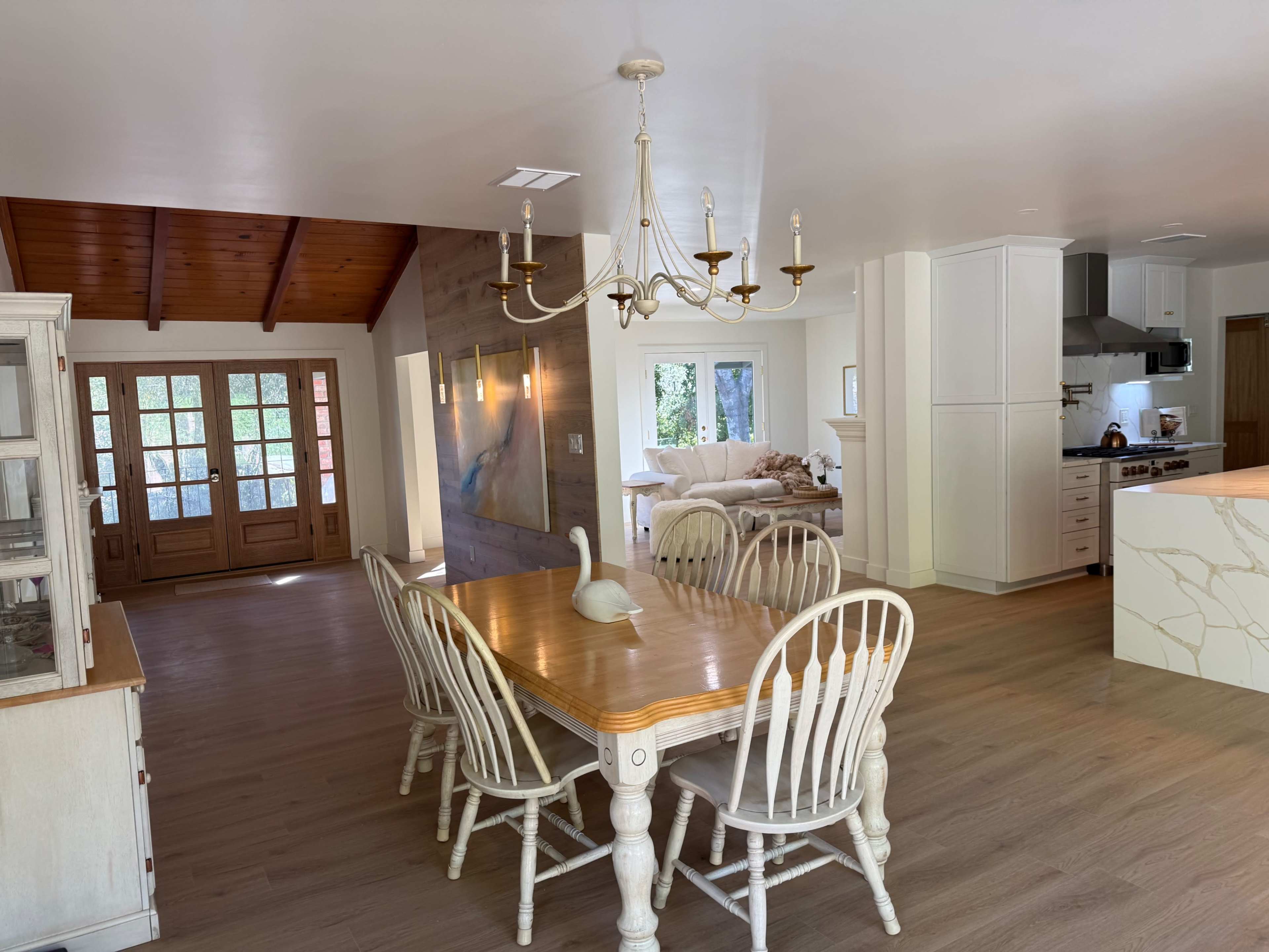 The image shows a spacious dining area with a wooden table and chairs, adjacent to a modern kitchen and a living room with large windows.