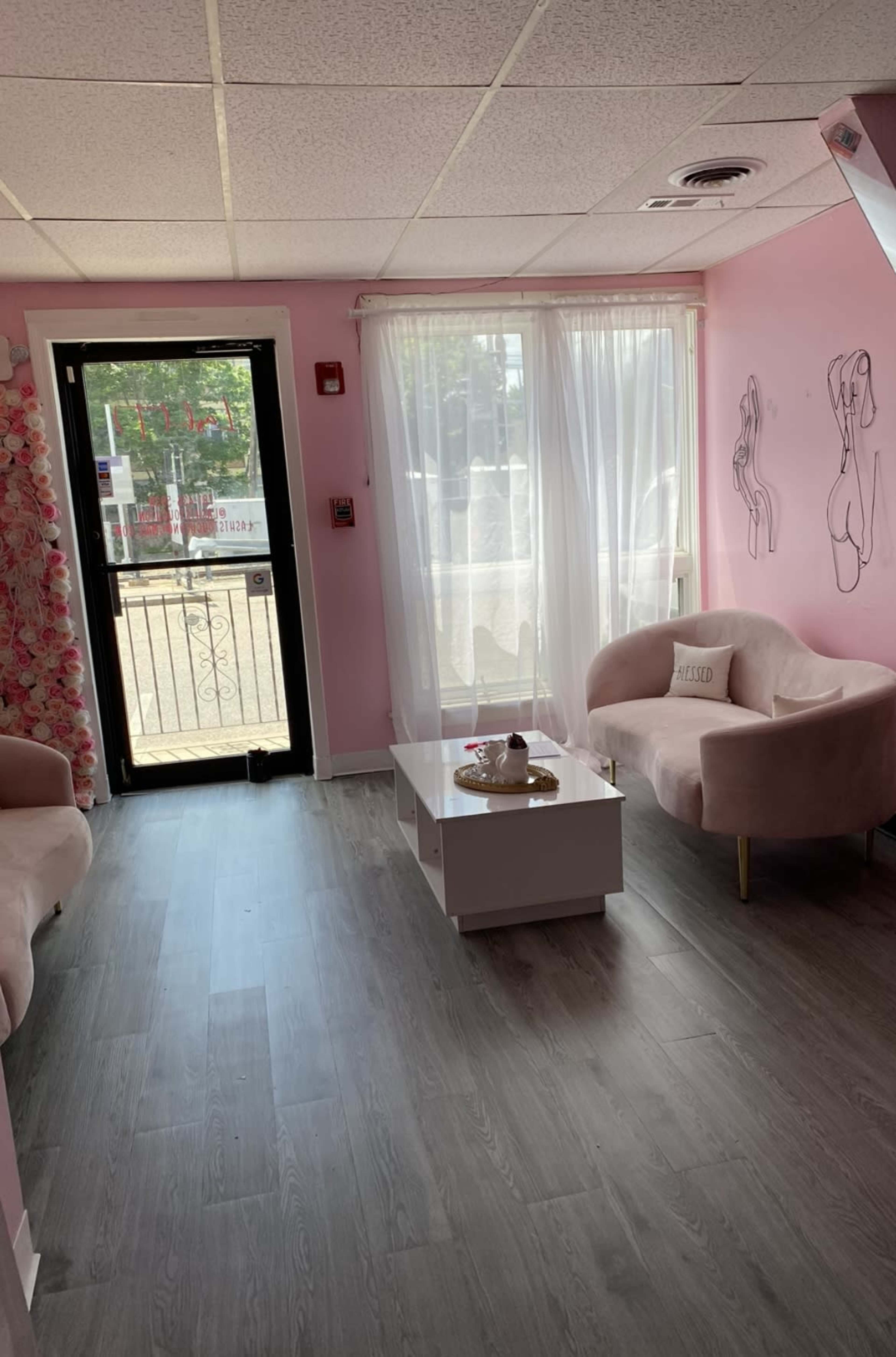 The image shows a brightly lit room with pink walls, a white window draped with sheer curtains, a small white coffee table, and two light pink chairs arranged around it.