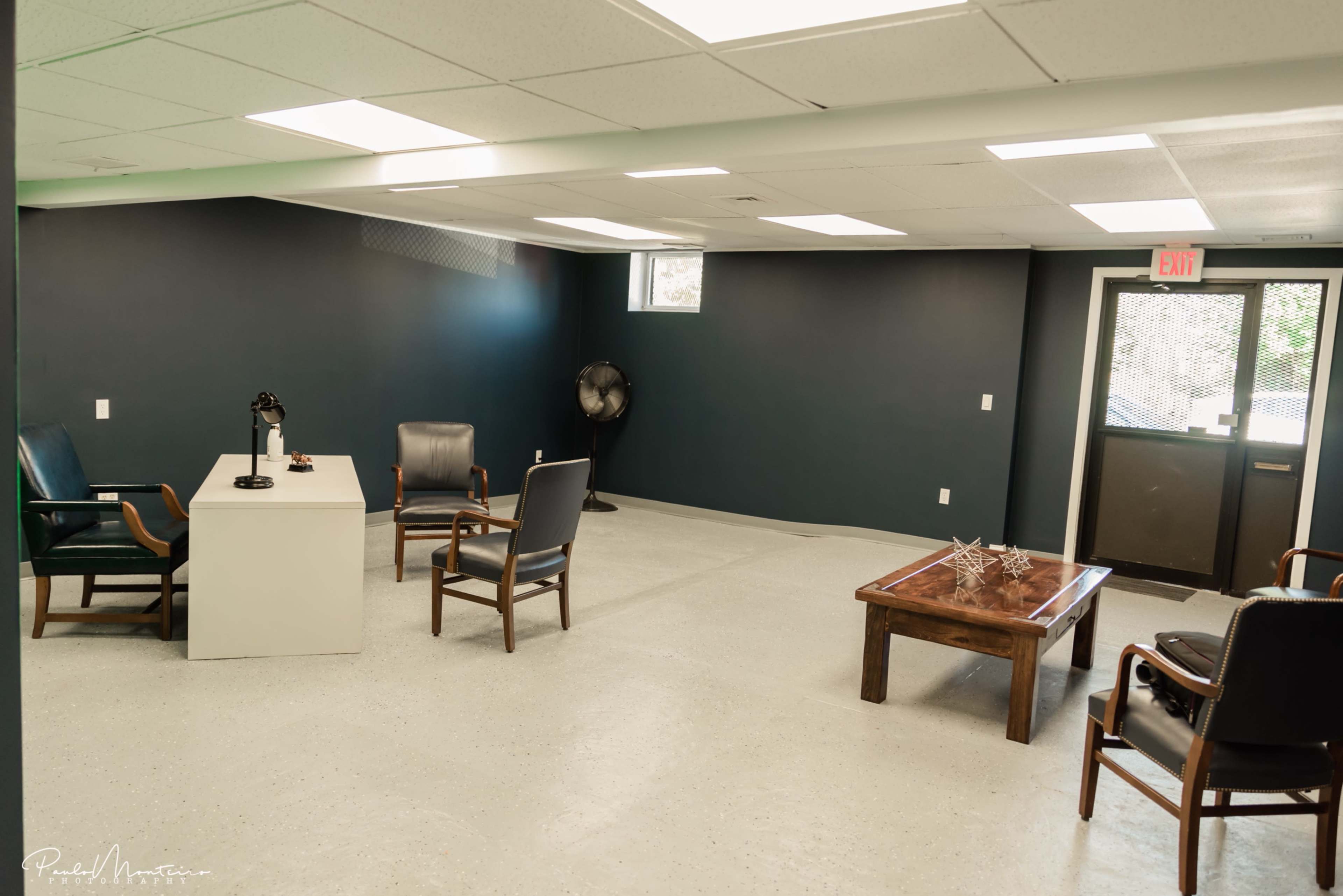 The image shows a sparsely furnished room with a white table, several chairs, and a large window.