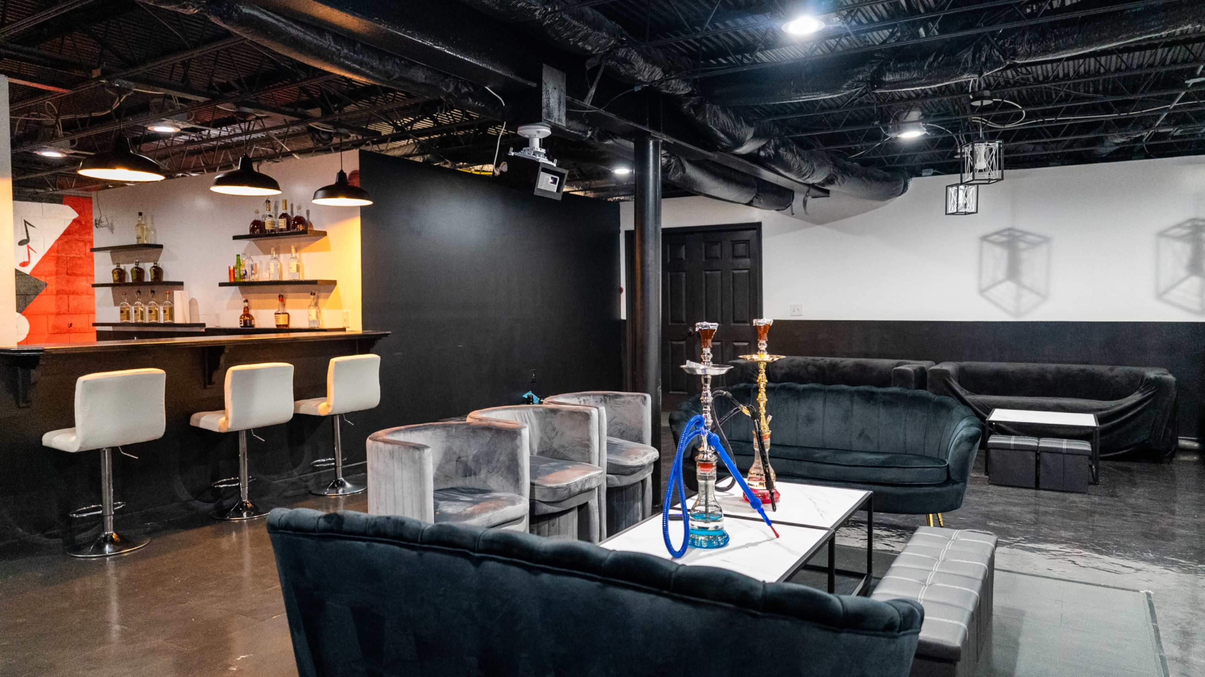 A modern lounge area featuring a bar with shelves of liquor, seating arrangements with plush couches, and shisha pipes on a table.