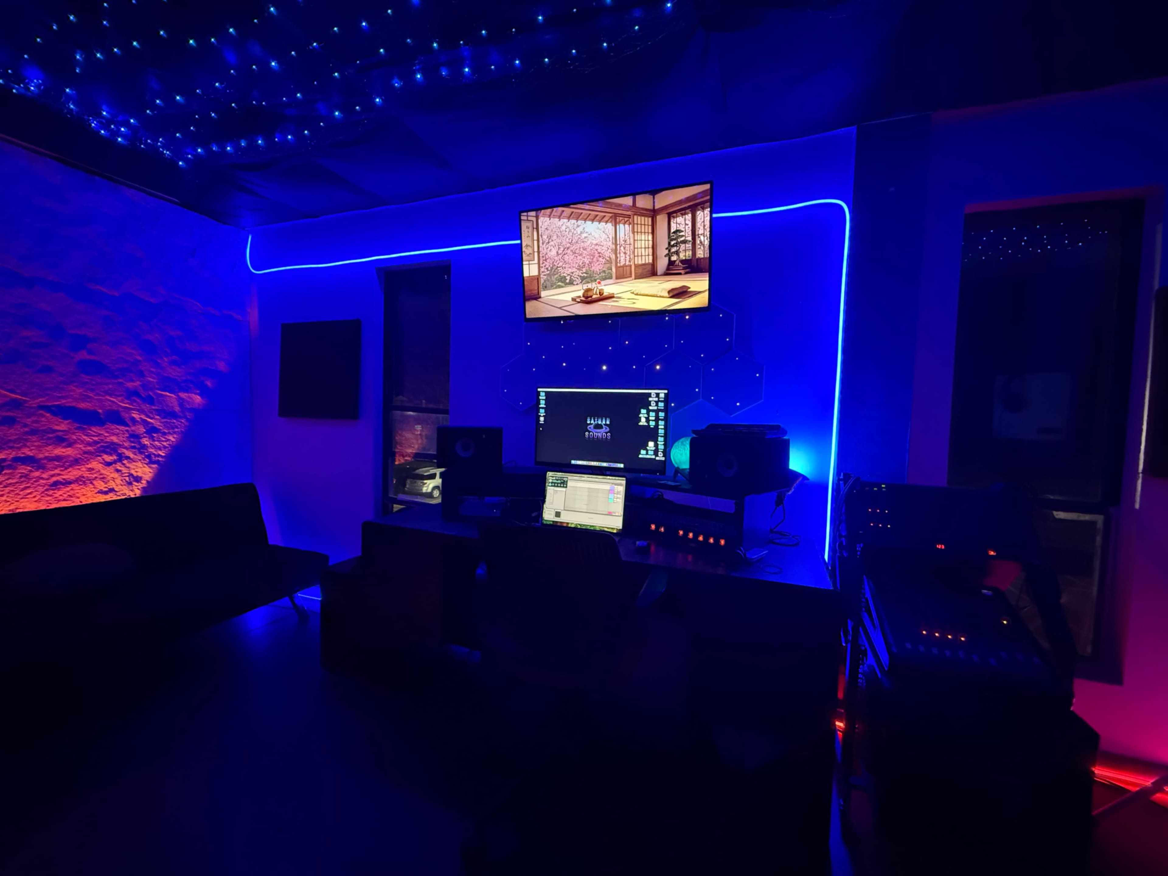 The image shows a dimly lit music studio with colorful LED lighting, featuring a computer setup, monitors, and a television screen on the wall.