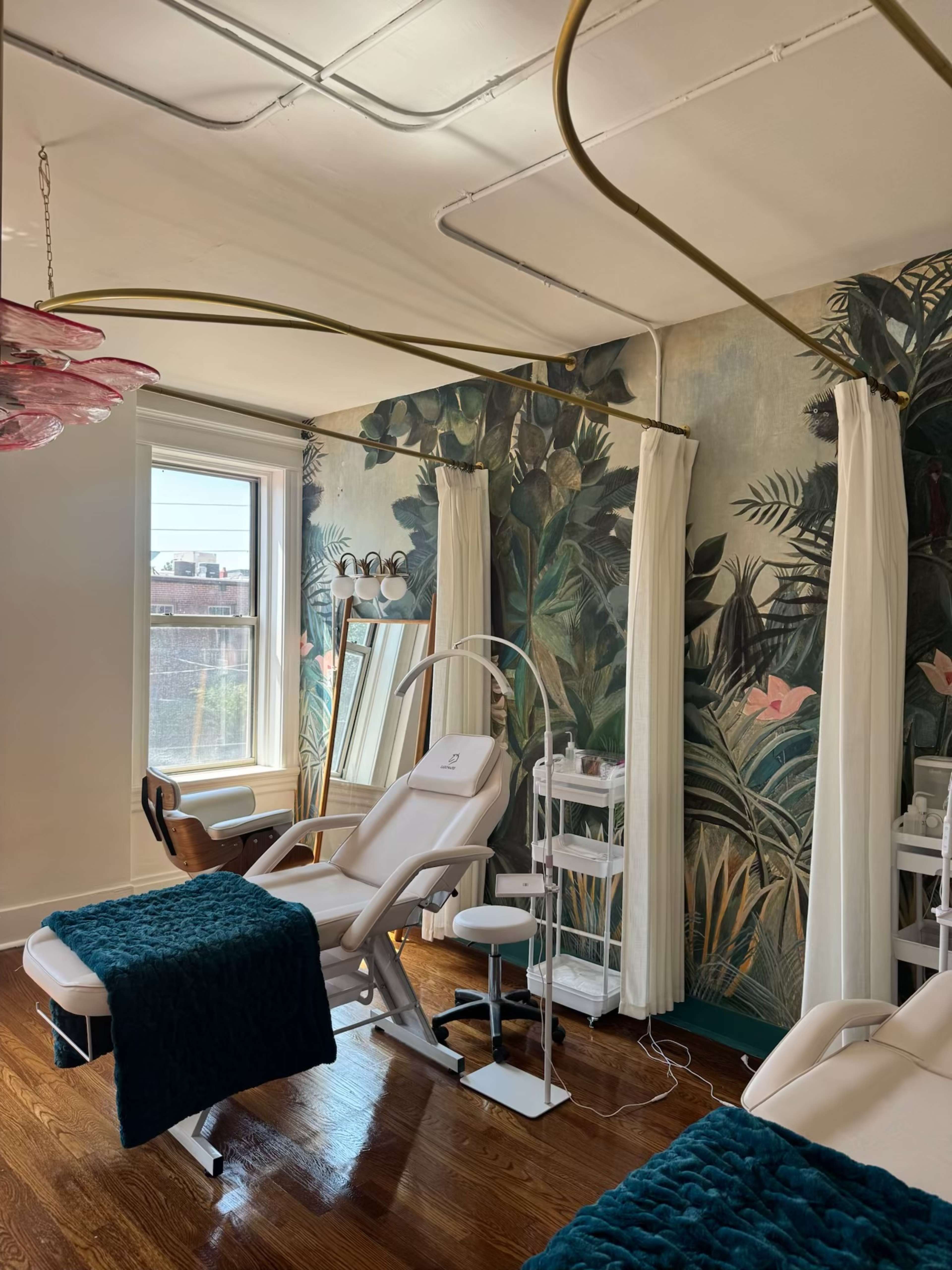 The image shows a spa room with two reclining treatment chairs, a standing lamp, and a decorative wall mural featuring tropical plants.