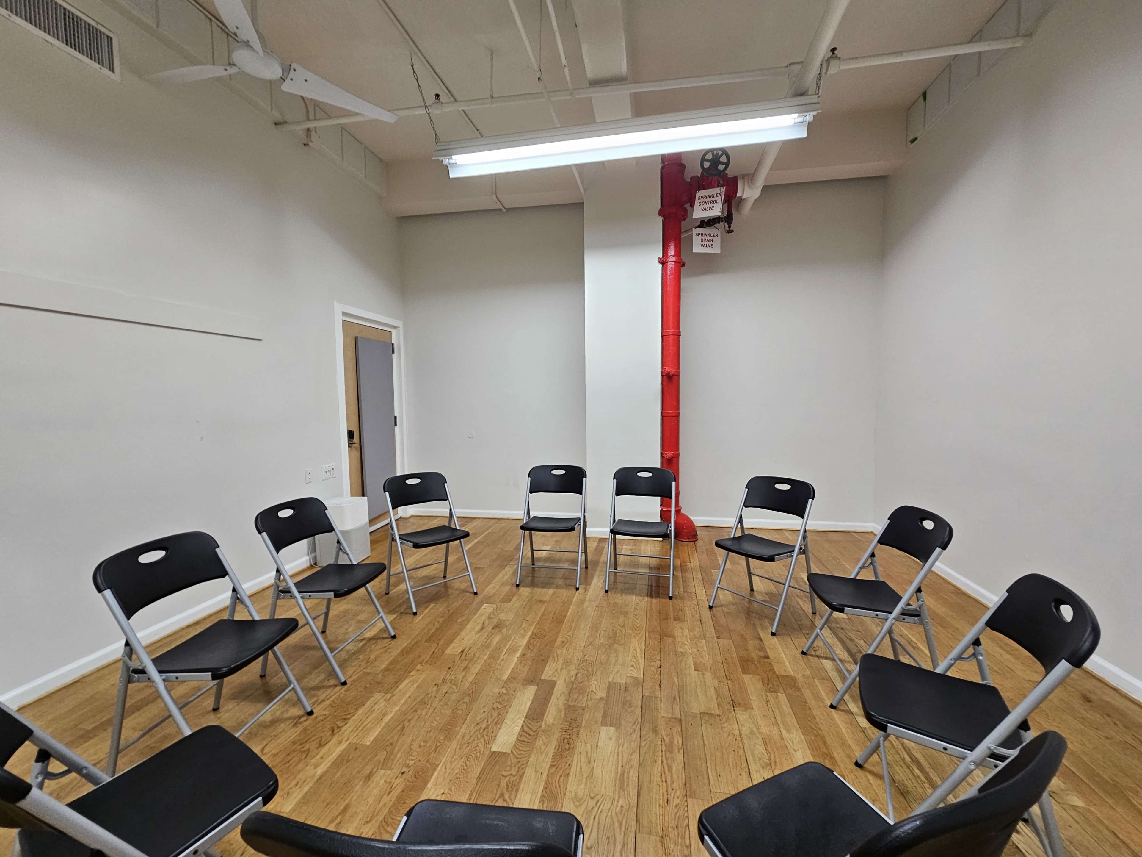 A room with a circular arrangement of black foldable chairs on a wooden floor, with a red pipe running along the wall.