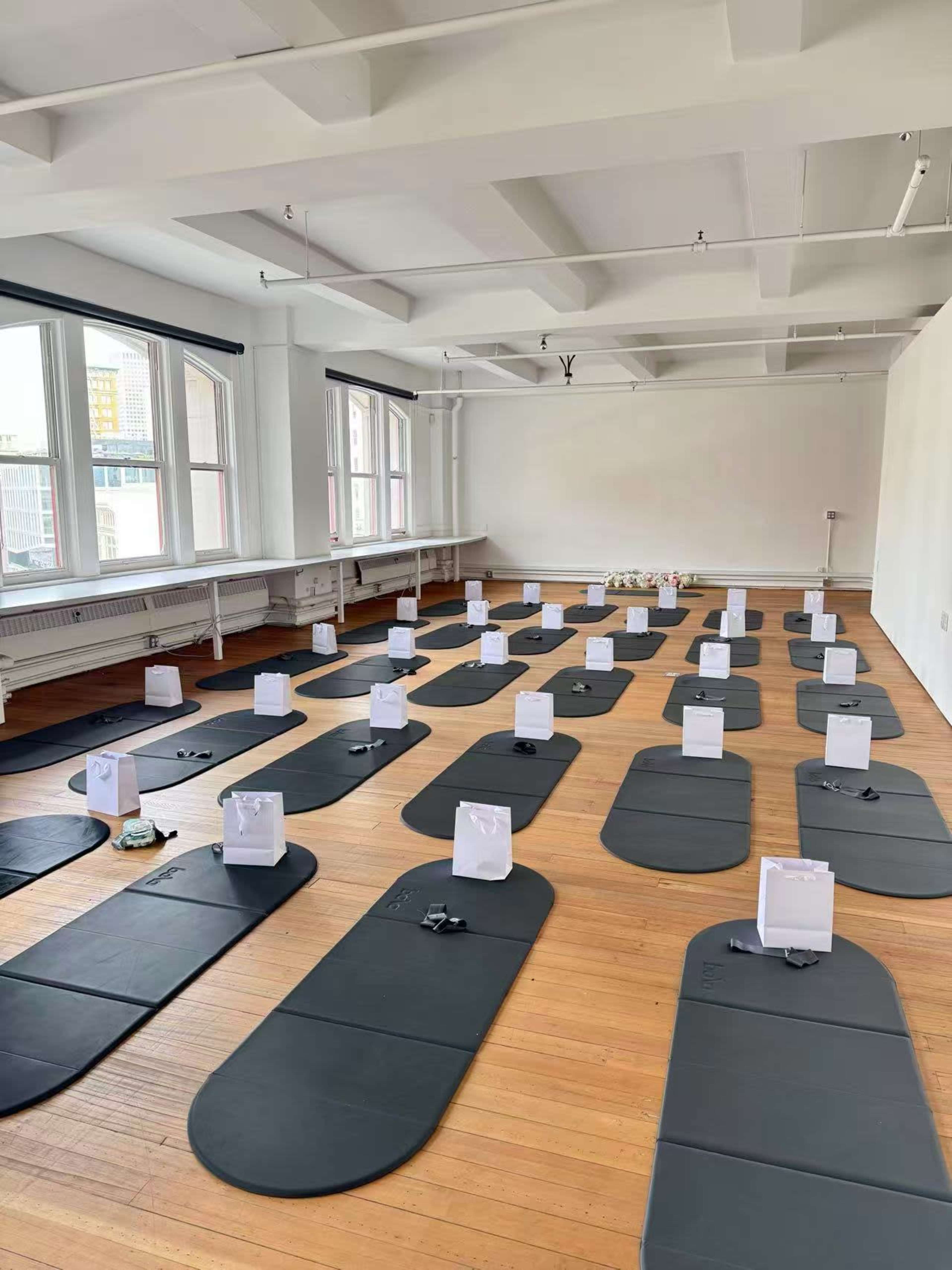 The image shows a spacious, brightly lit room arranged with several black yoga mats, each accompanied by a white item placed on them, likely for a yoga or meditation session.