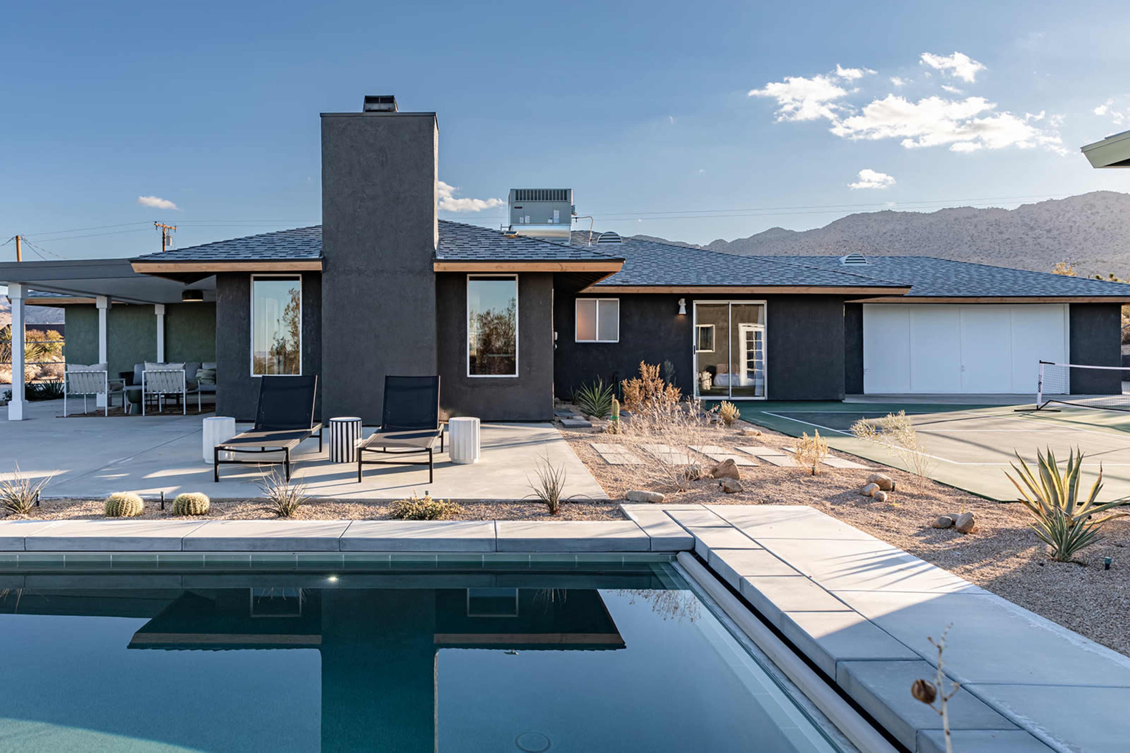 A modern house with a pool is set against a mountainous backdrop, featuring a sleek design and outdoor furniture.