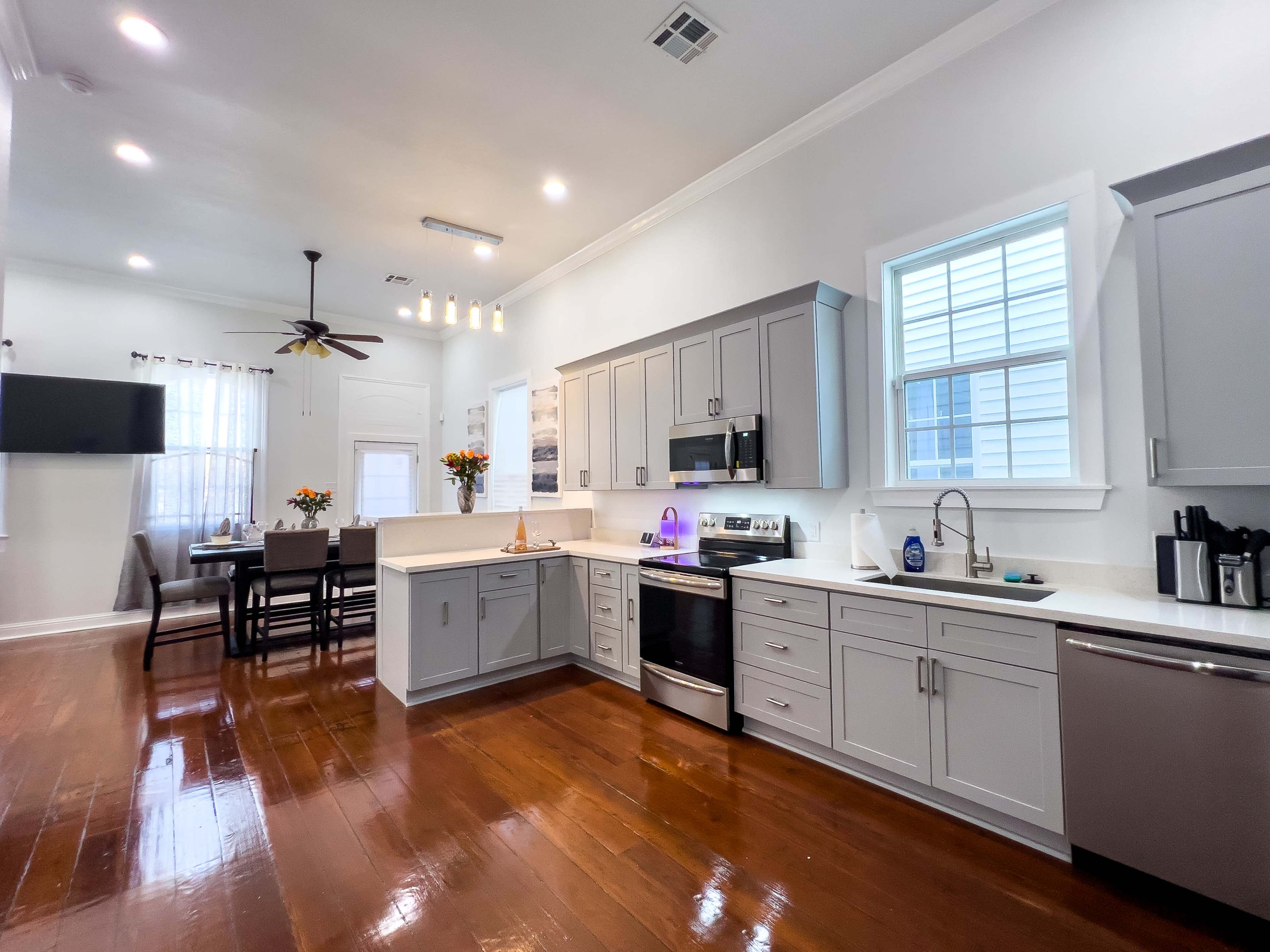 The image shows a modern kitchen with gray cabinets, stainless steel appliances, and a dining area featuring a table and chairs in a well-lit space.