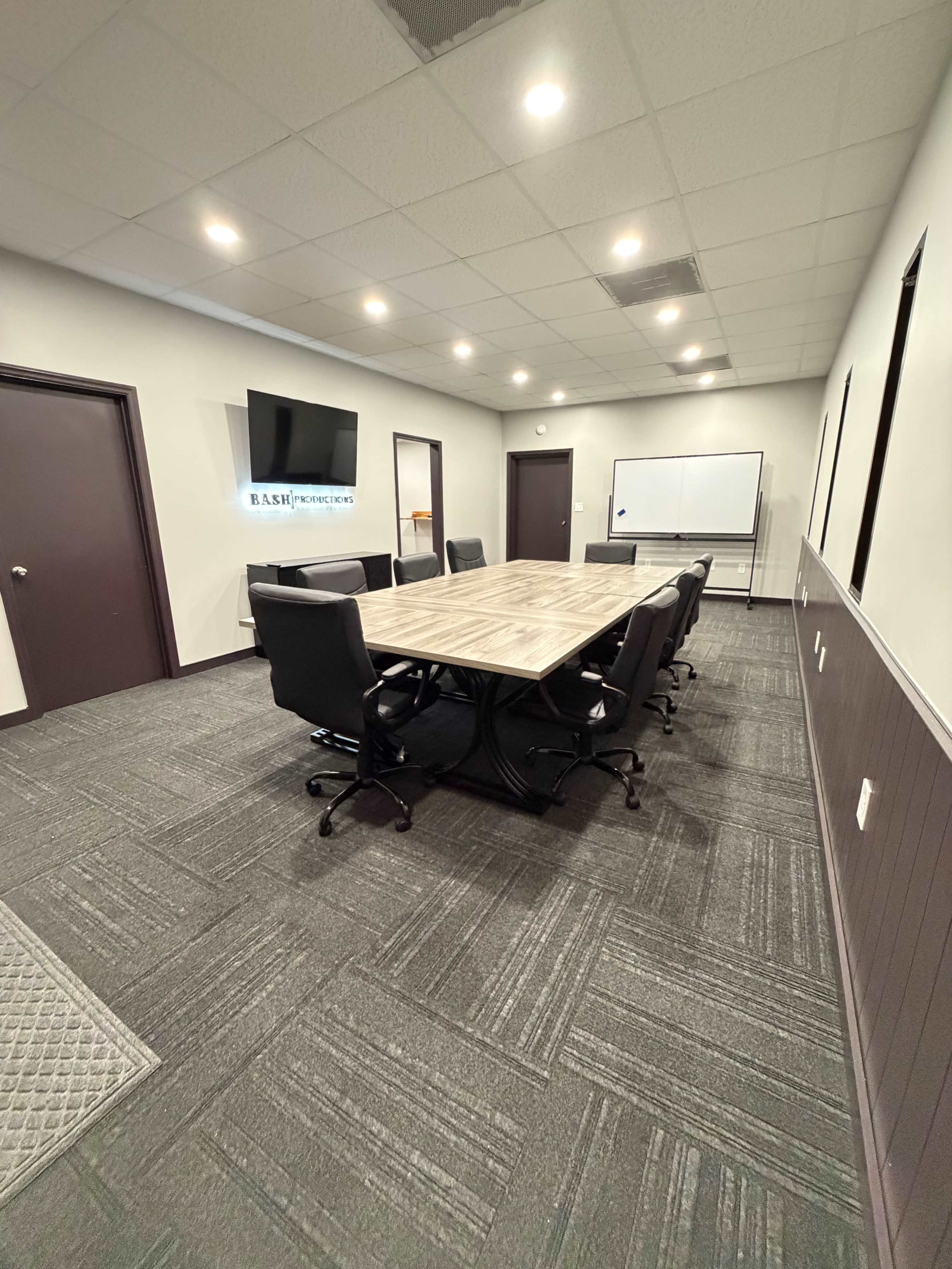 The image shows a spacious conference room with a large rectangular table surrounded by black chairs, featuring a mounted TV and a whiteboard on the walls.