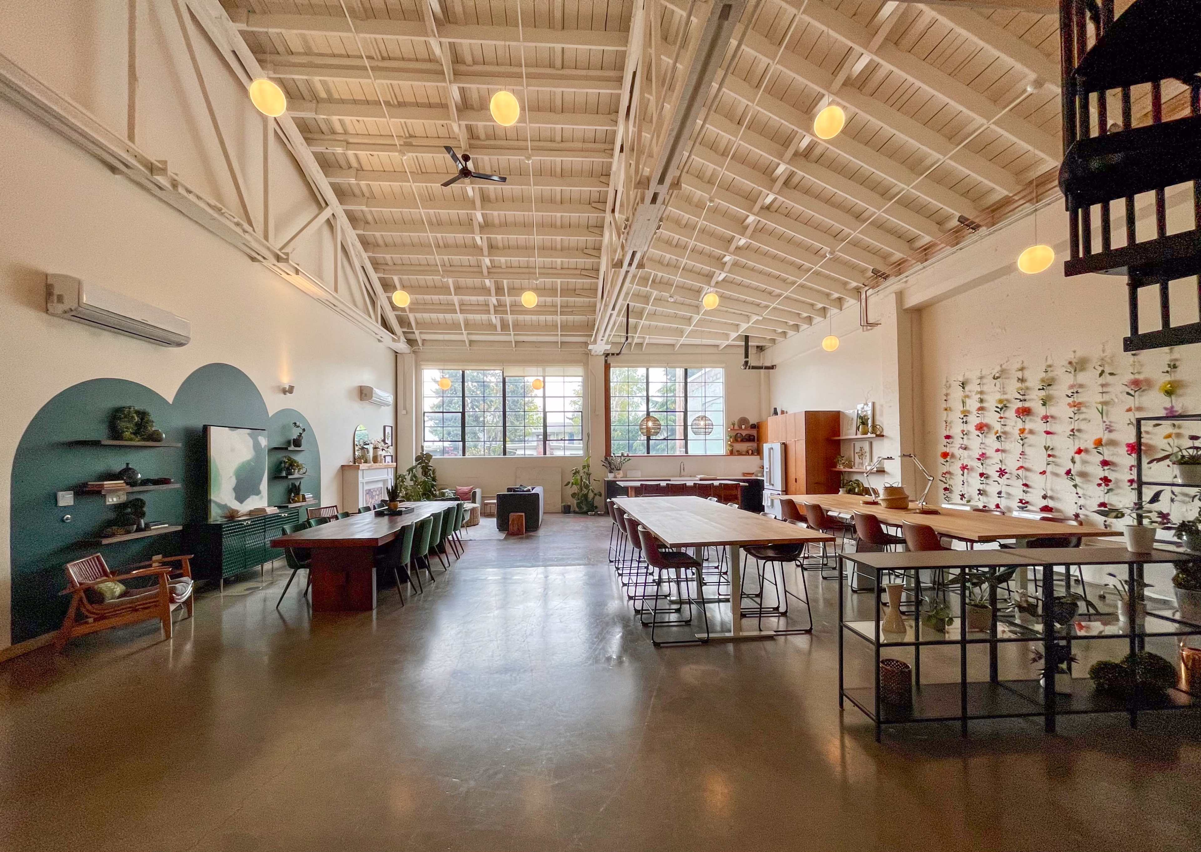 The image shows a spacious room with high ceilings, featuring large windows, a mix of dining and work tables, and a vertical garden on one wall.