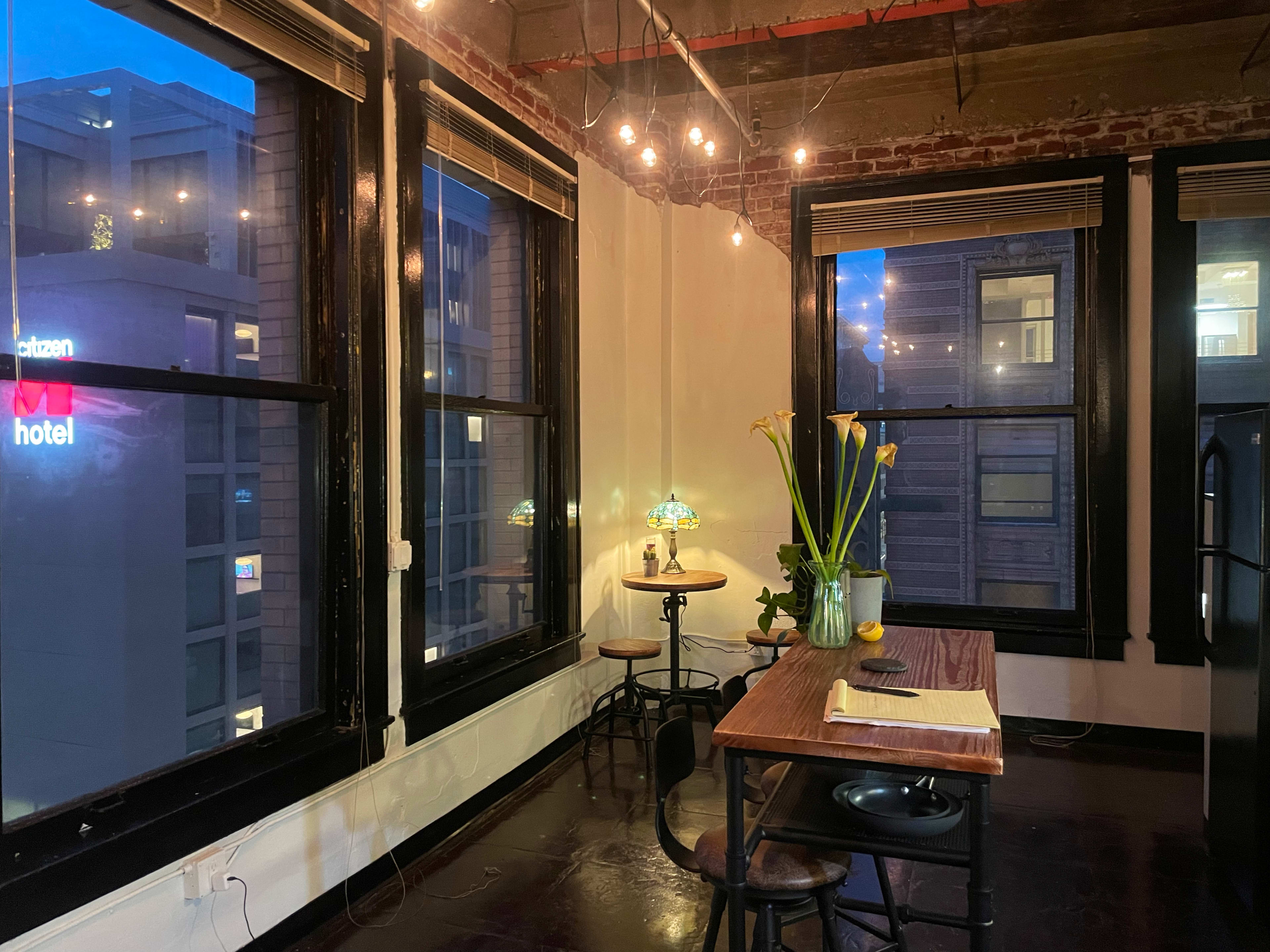 A cozy interior space with a wooden table and chairs, large windows displaying city lights outside, and a vase of flowers on the table.