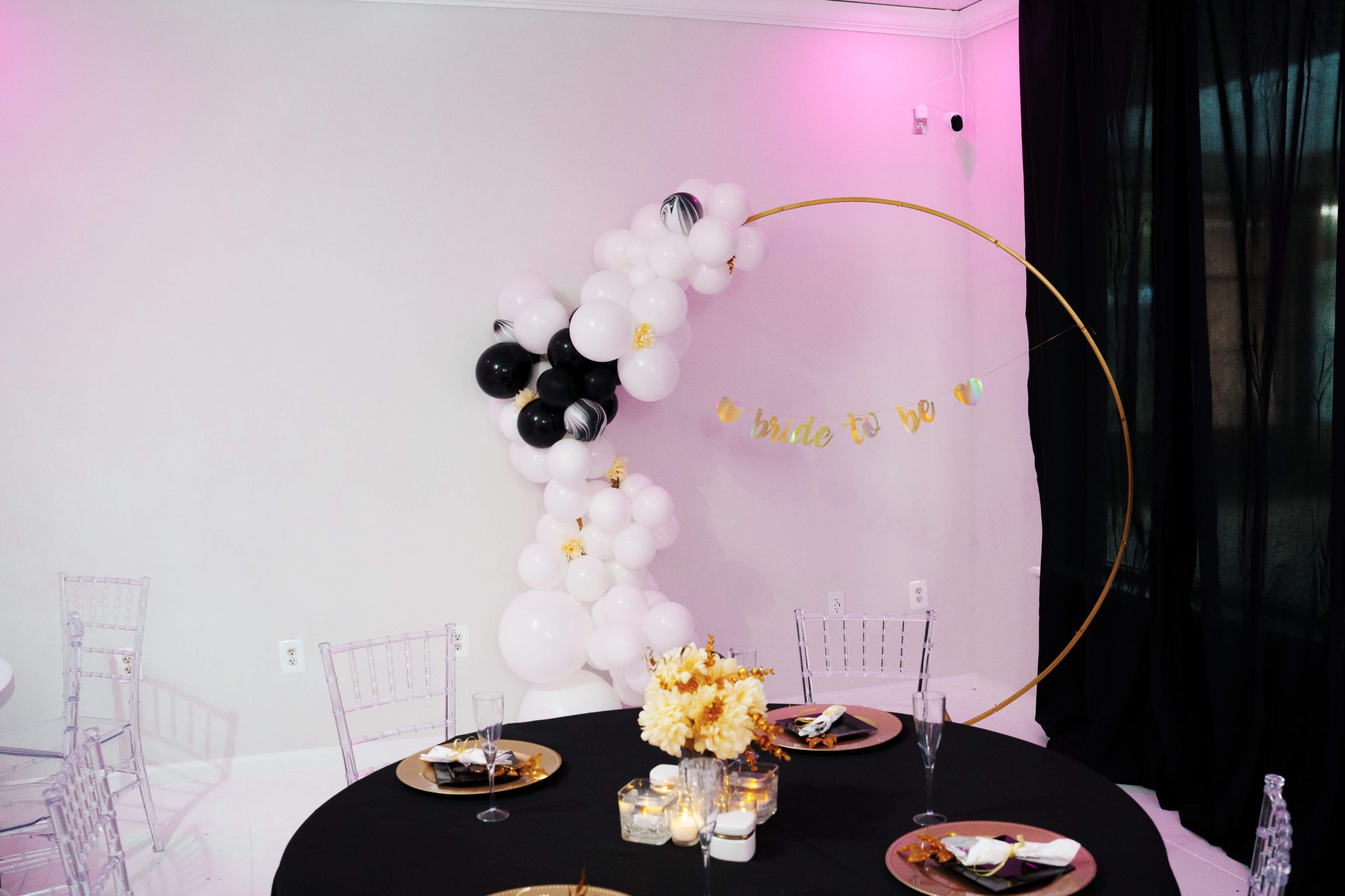 The scene features a decorated table setting with black and gold accents, a backdrop of balloon arrangements, and a "bride to be" banner in a celebration space.