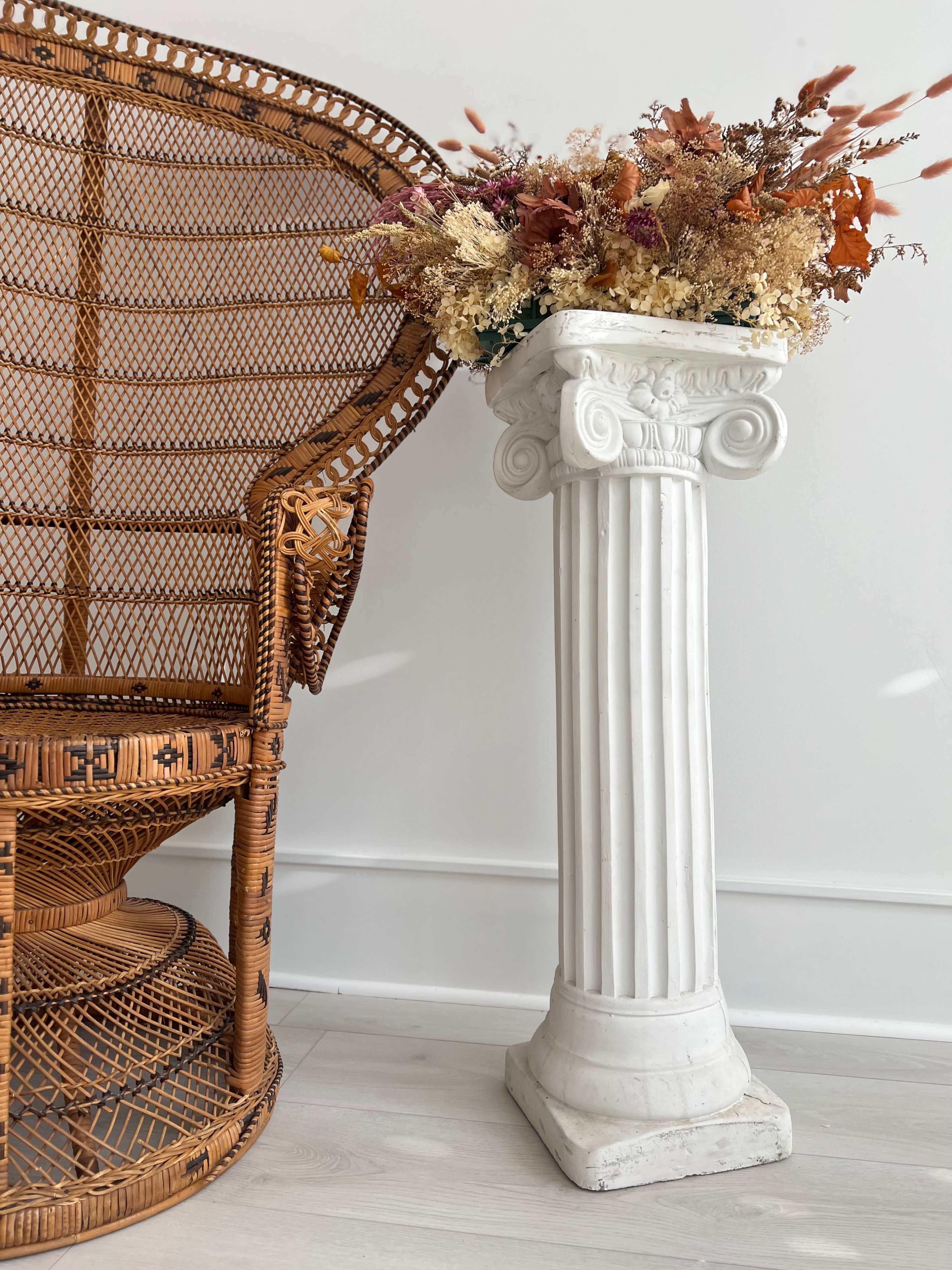 A wicker chair is positioned next to a white column base that supports a decorative arrangement of dried flowers.