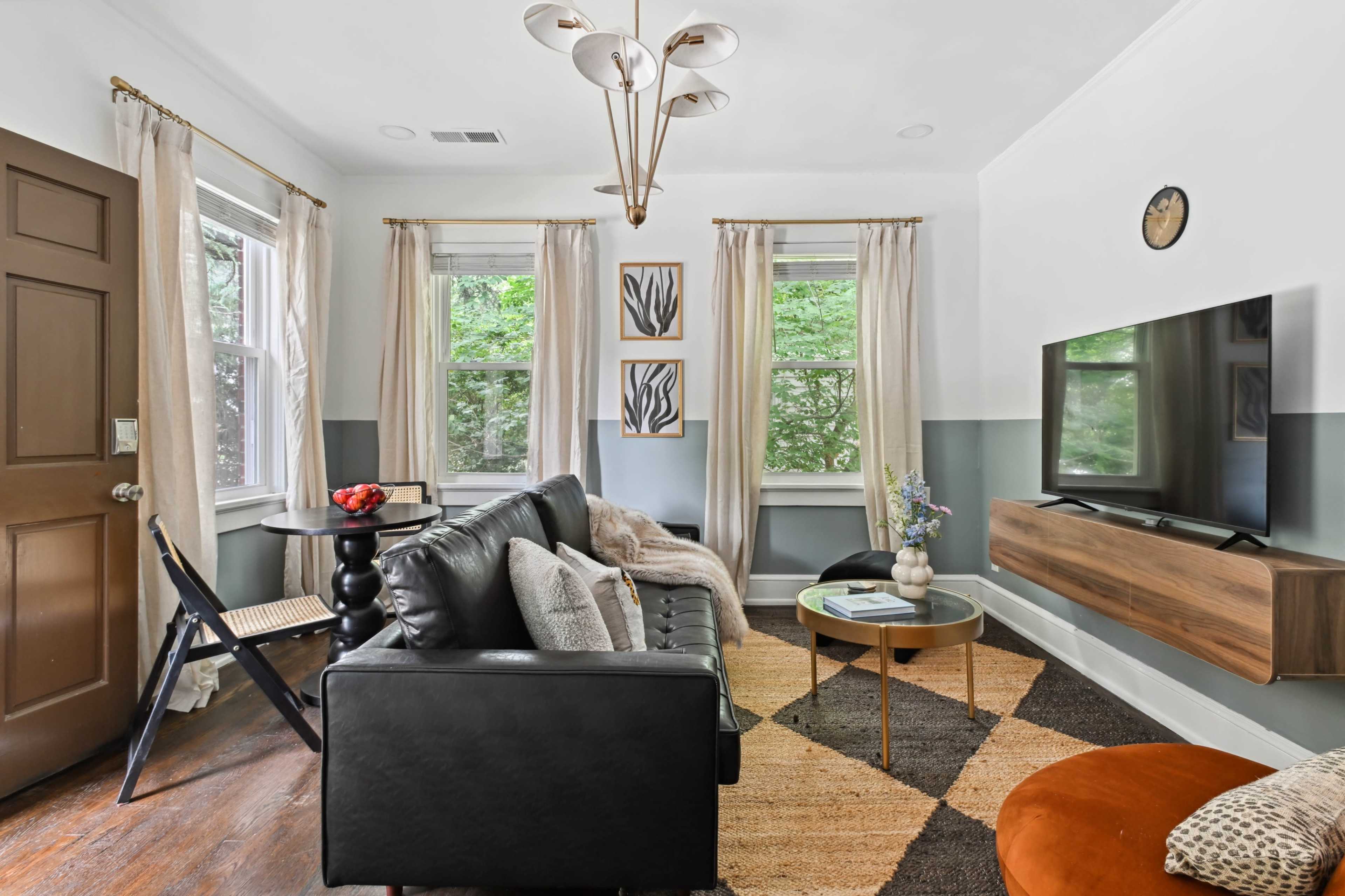 The living room features a black leather sofa, a wooden TV stand, a round coffee table, and large windows with light-colored curtains.