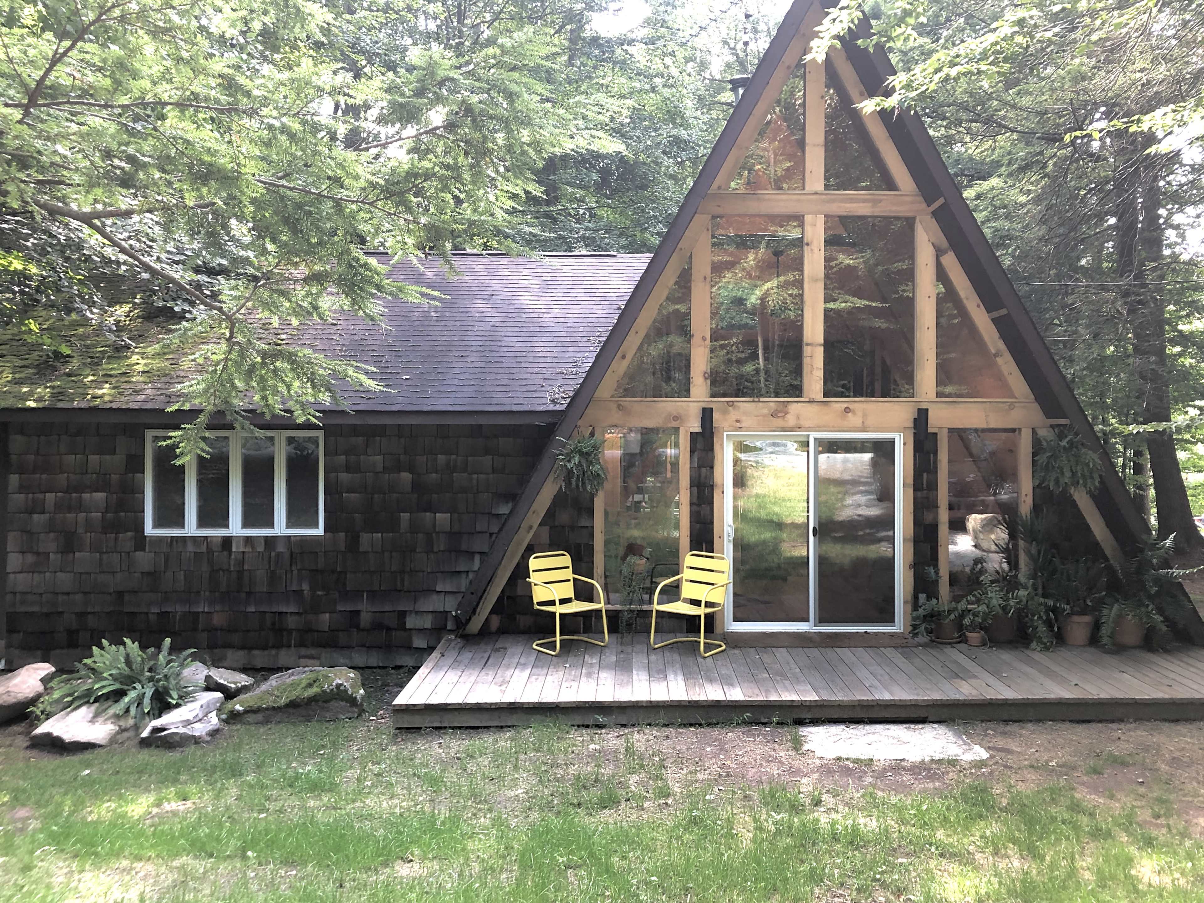 A wooden A-frame cabin with large front windows features a small deck and two yellow chairs.