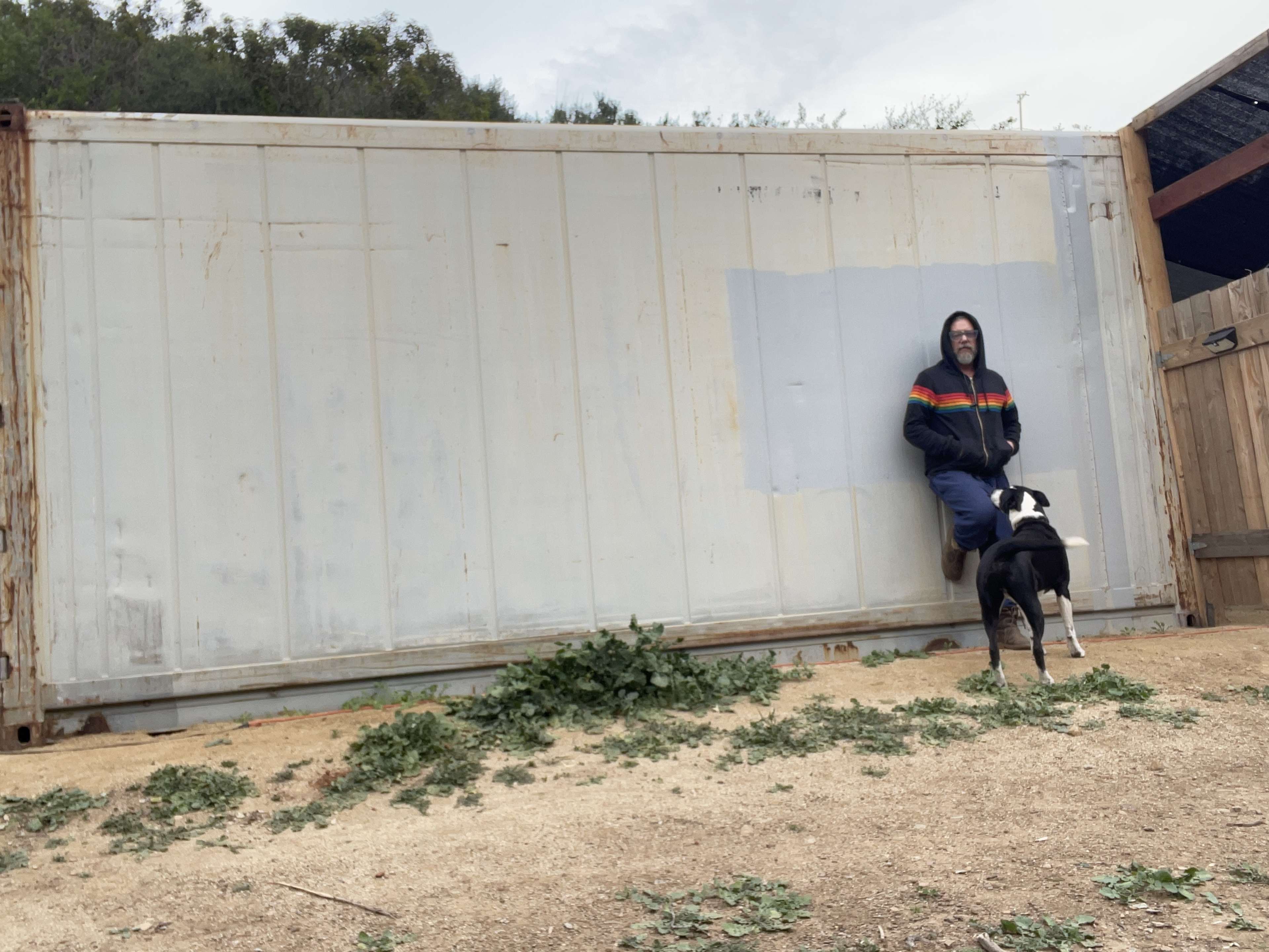 A person sits against a shipping container while a dog stands nearby on a dirt surface with scattered vegetation.