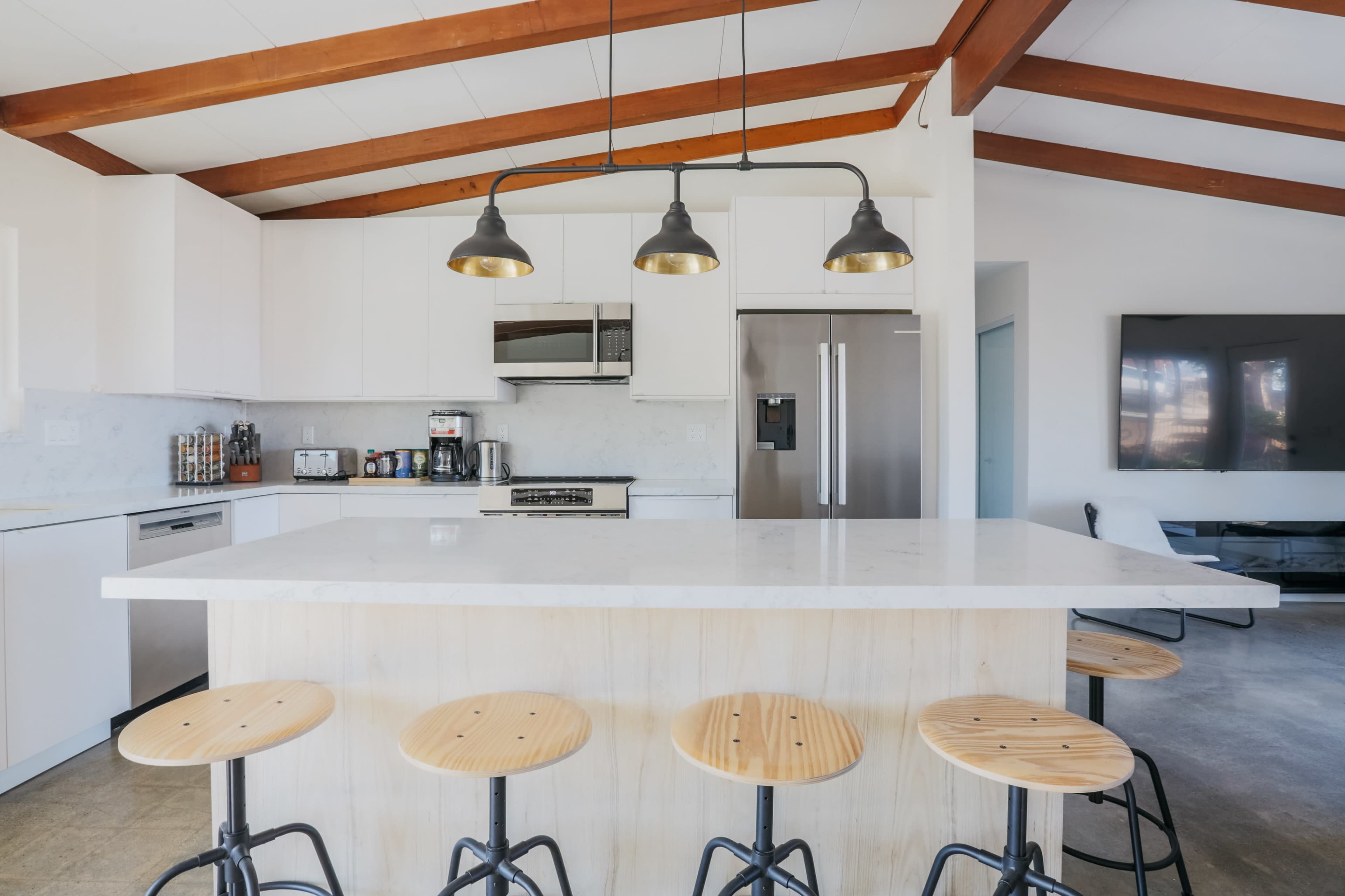 A modern kitchen features a large island with wooden stools and overhead pendant lights, set against white cabinetry and a stainless steel refrigerator.