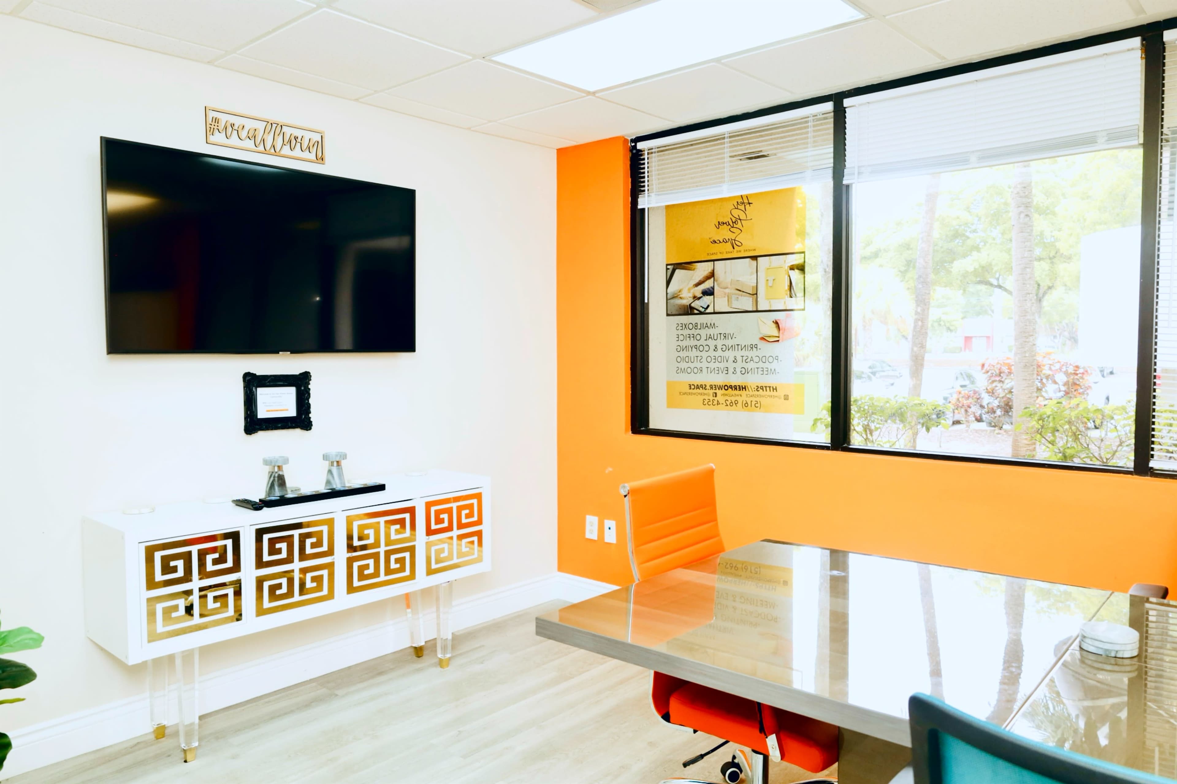 The image shows a modern office space with an orange accent wall, a sleek white cabinet with gold detailing, a television mounted on the wall, and a glass-top table surrounded by orange and white chairs.