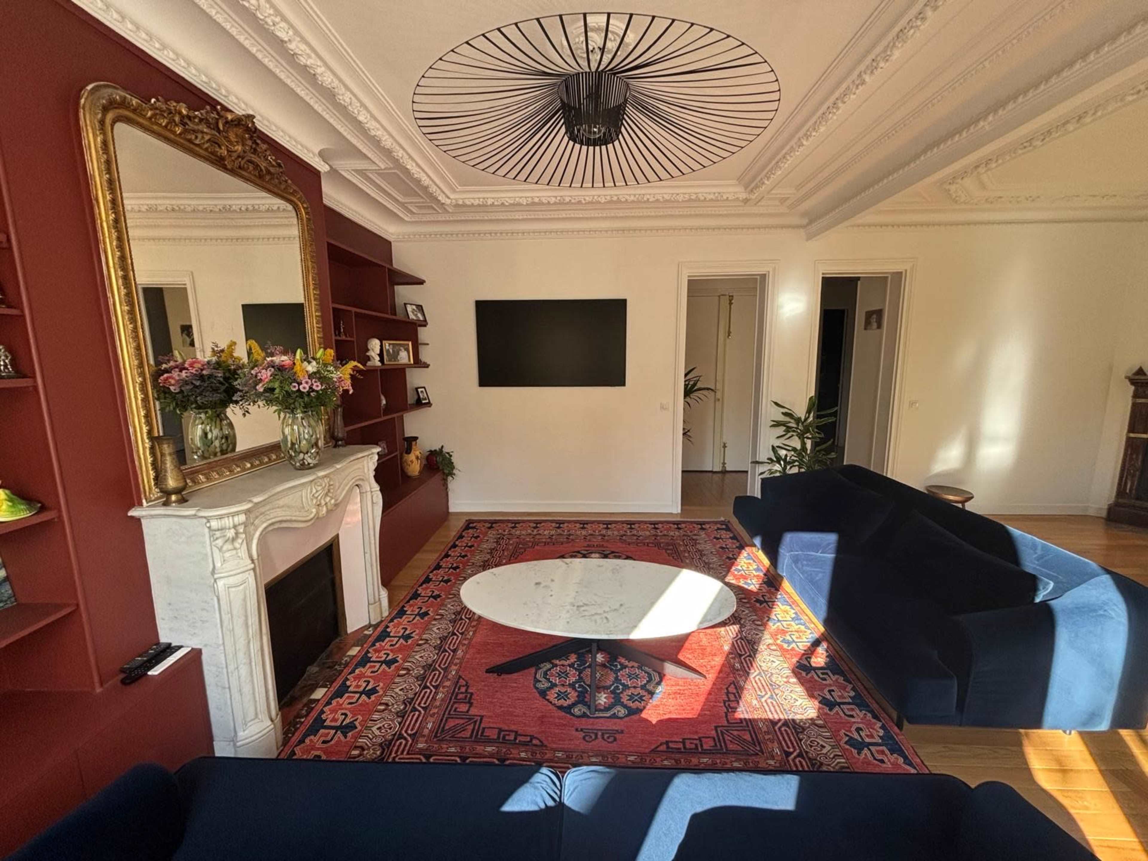 A cozy living room features two blue sofas, a circular marble coffee table, a decorative fireplace with a mirror above it, and a wall-mounted television against a red accent wall.
