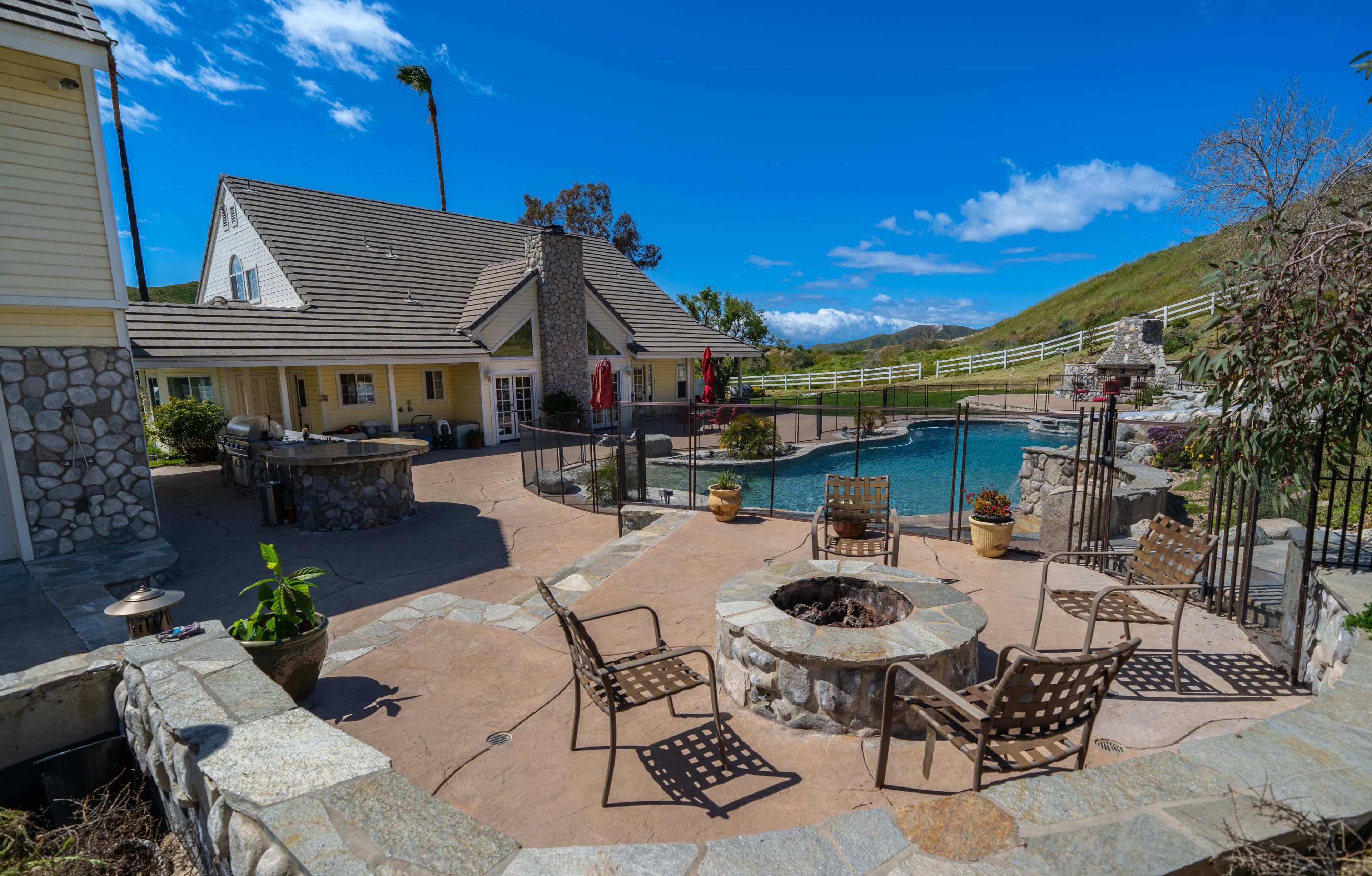 A spacious outdoor area features a stone fire pit surrounded by metal chairs, a swimming pool, and a large house with a stone and wood exterior.