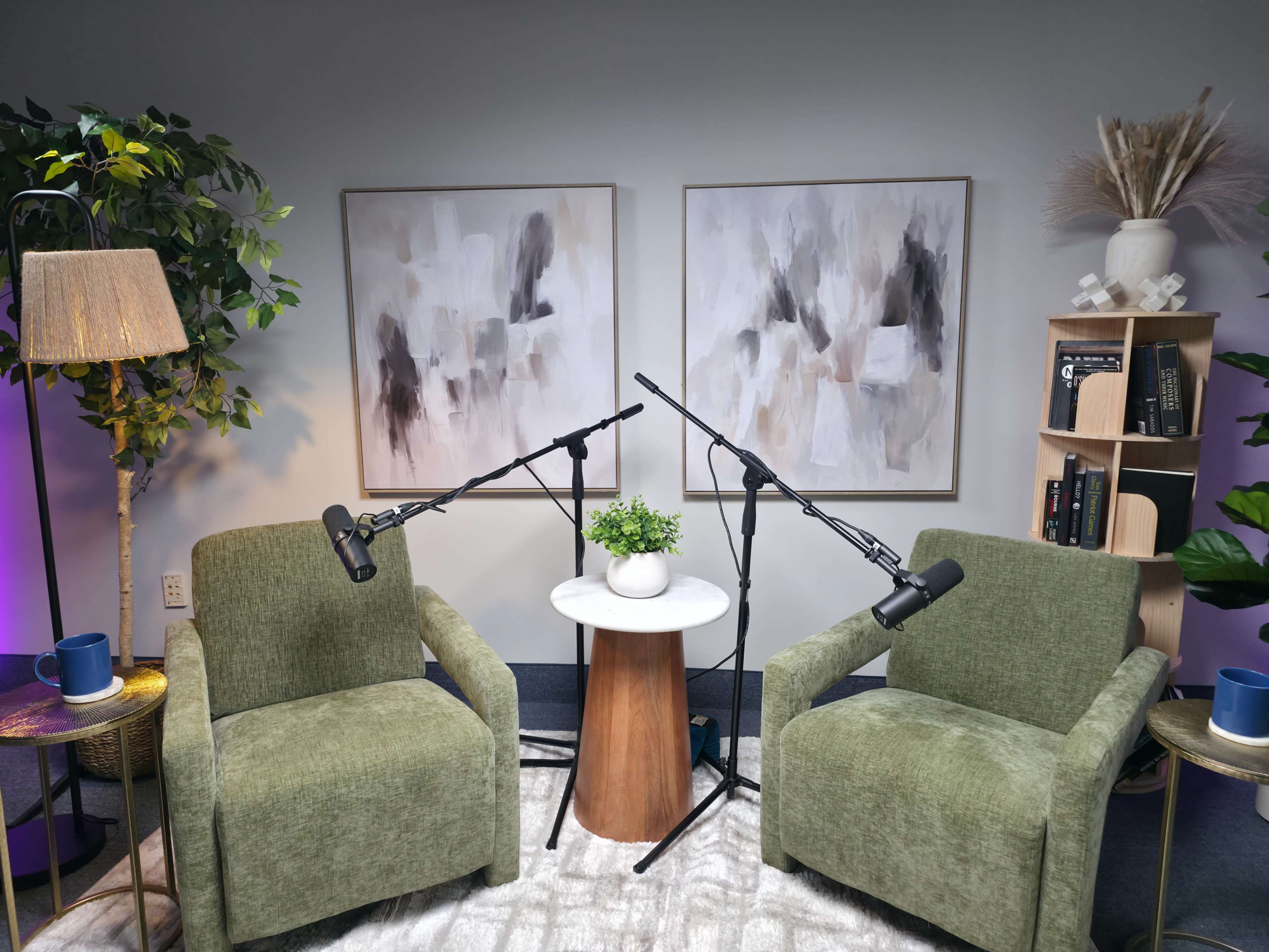 The image shows a cozy interview setup with two green chairs, a small round table, and microphones positioned on stands between them, surrounded by decorative plants and artwork on the walls.