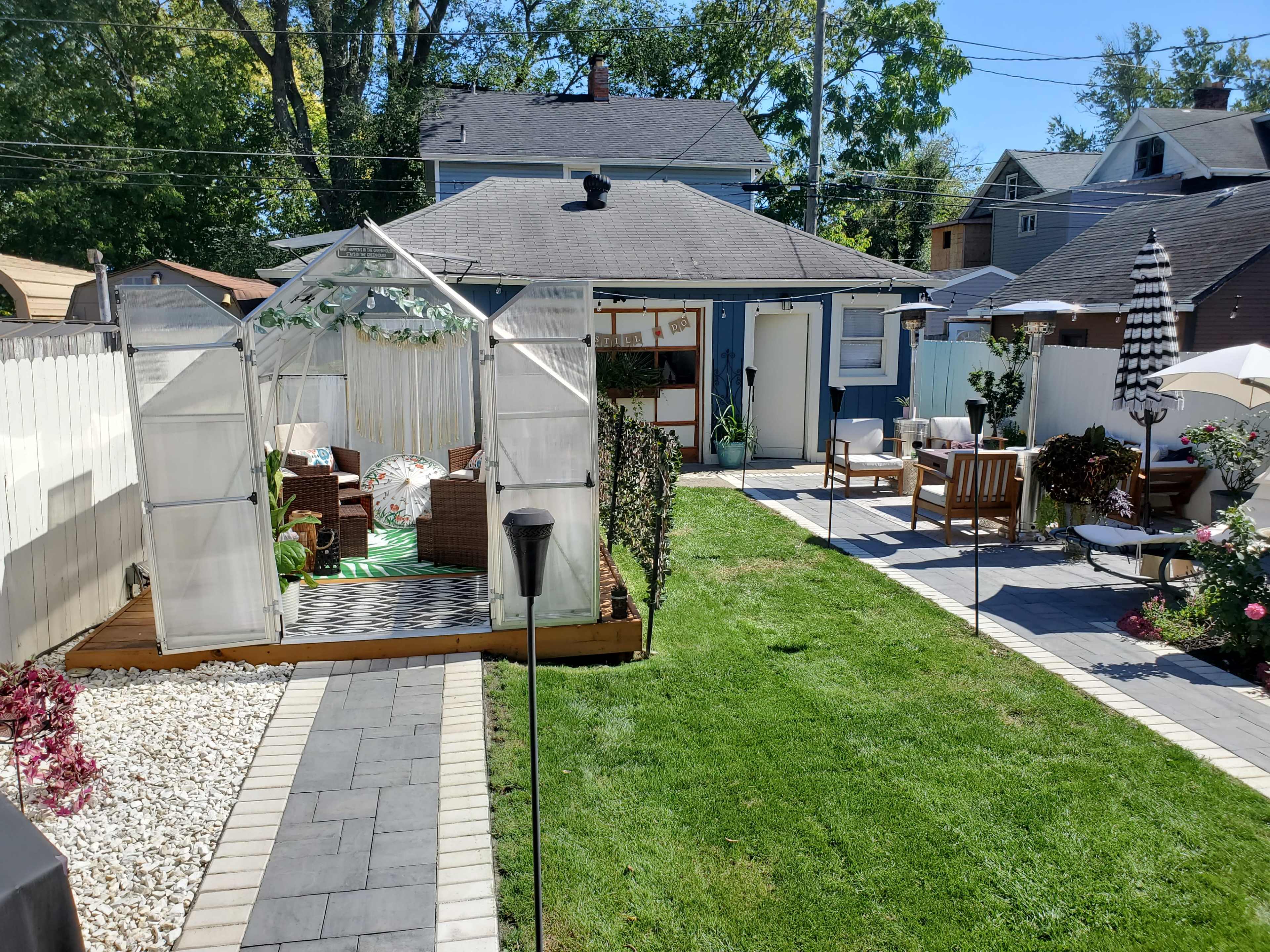 A landscaped backyard featuring a greenhouse, patio furniture, and pathways bordered by grass and decorative plants.