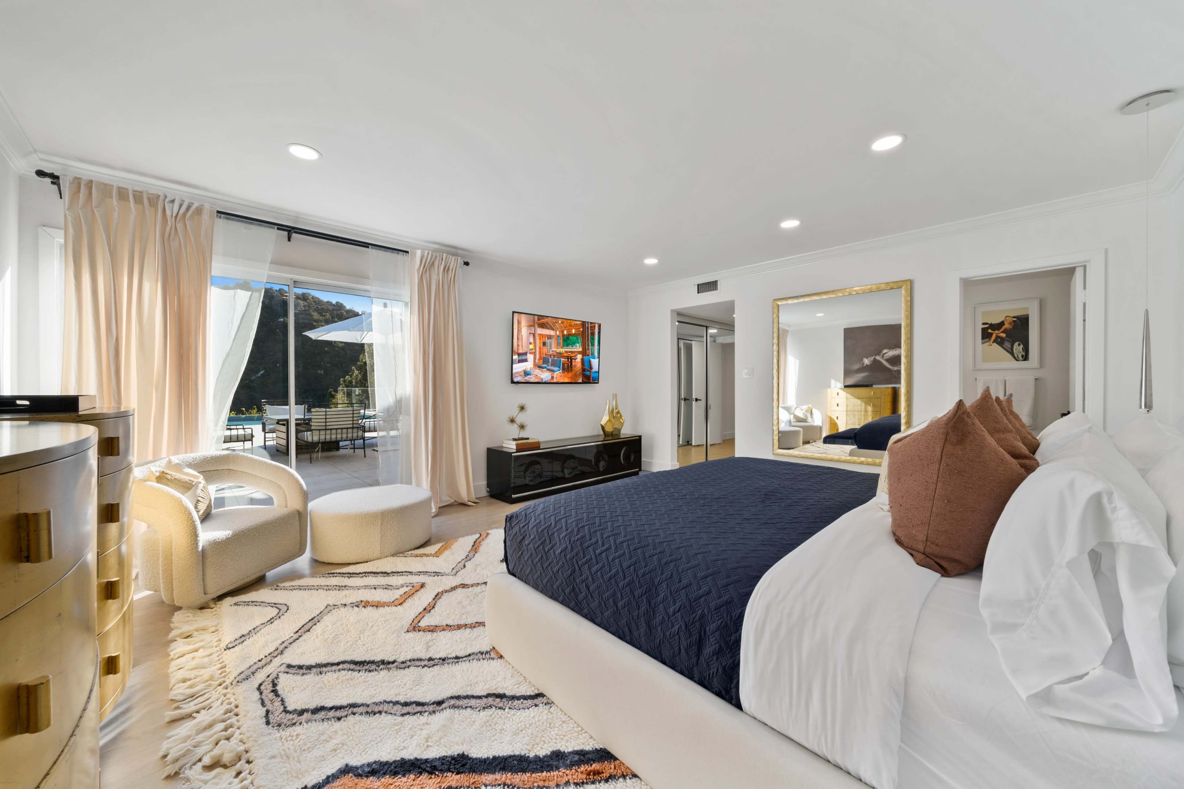 A bright bedroom features a large bed, a seating area with a chair and ottoman, a television mounted on the wall, a mirrored dresser, and a sliding door leading to a balcony.