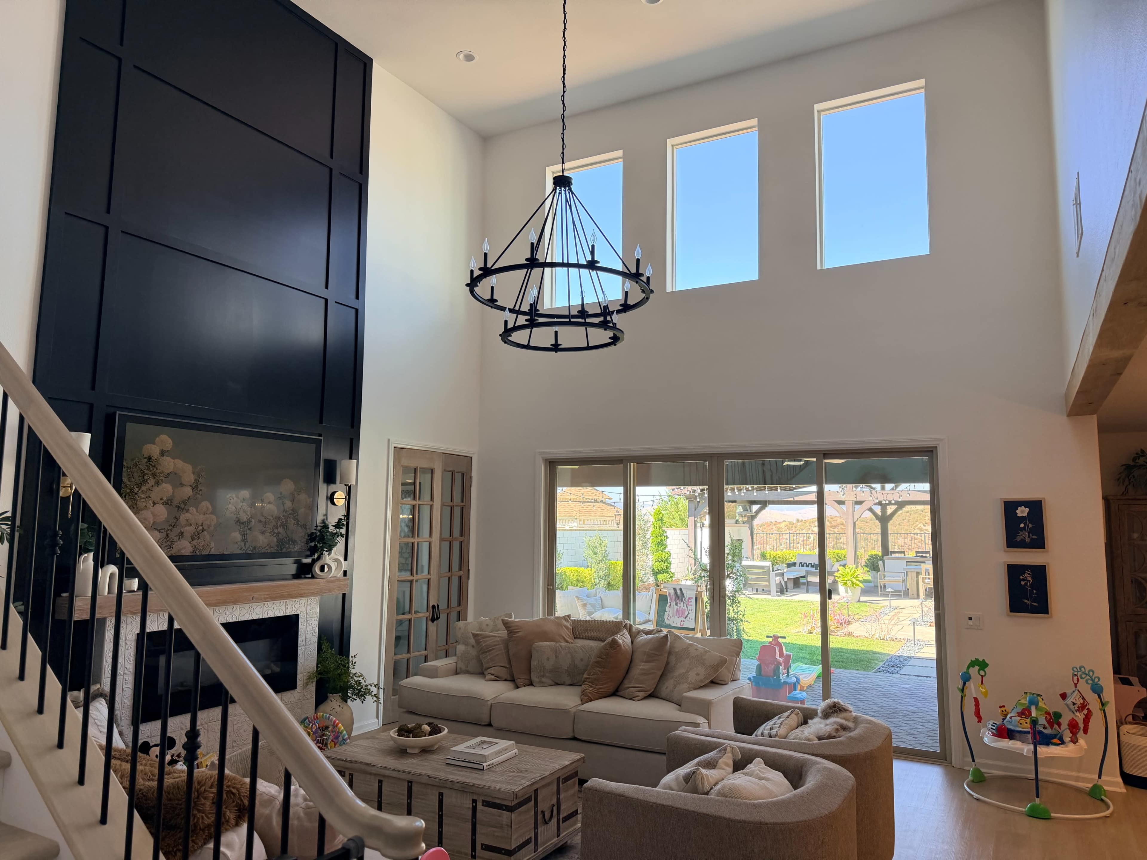The image shows a spacious living room with high ceilings, large windows allowing natural light, and a view of an outdoor patio and garden area.