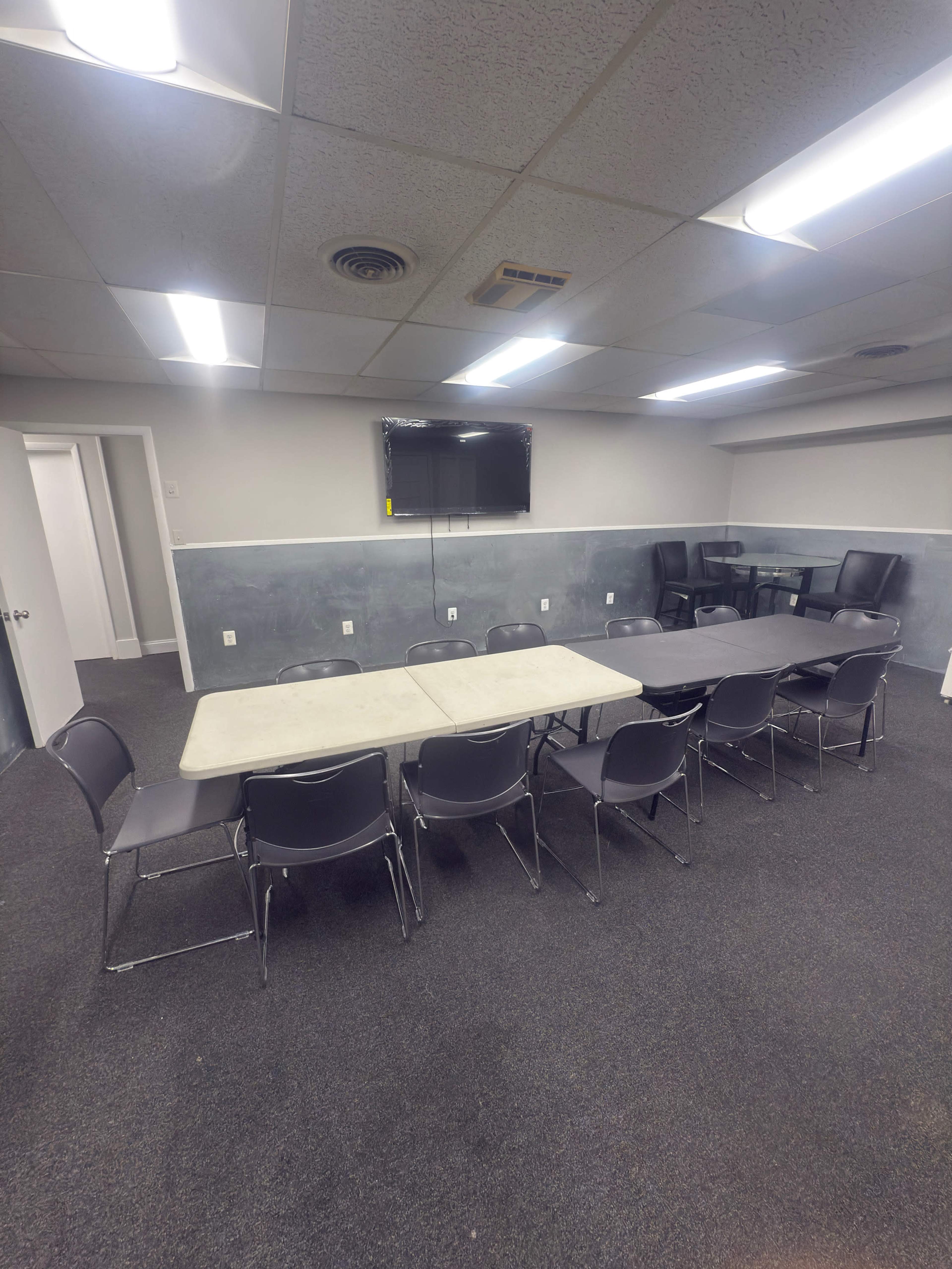 The image shows a meeting room with a large table surrounded by chairs, a television mounted on the wall, and another table in the background.