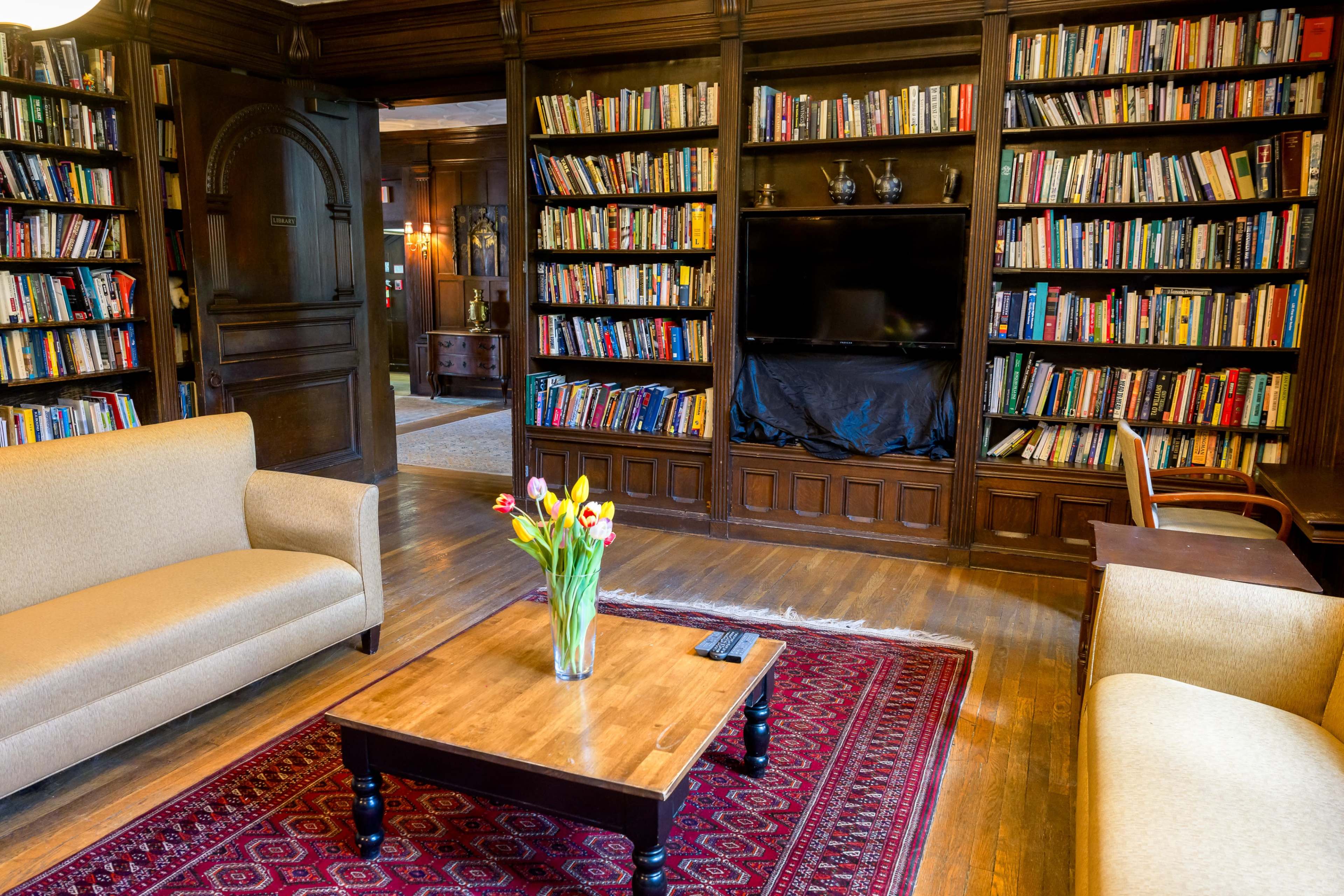 A cozy library featuring two beige sofas facing a wooden table, surrounded by bookshelves filled with books and a flat-screen television.