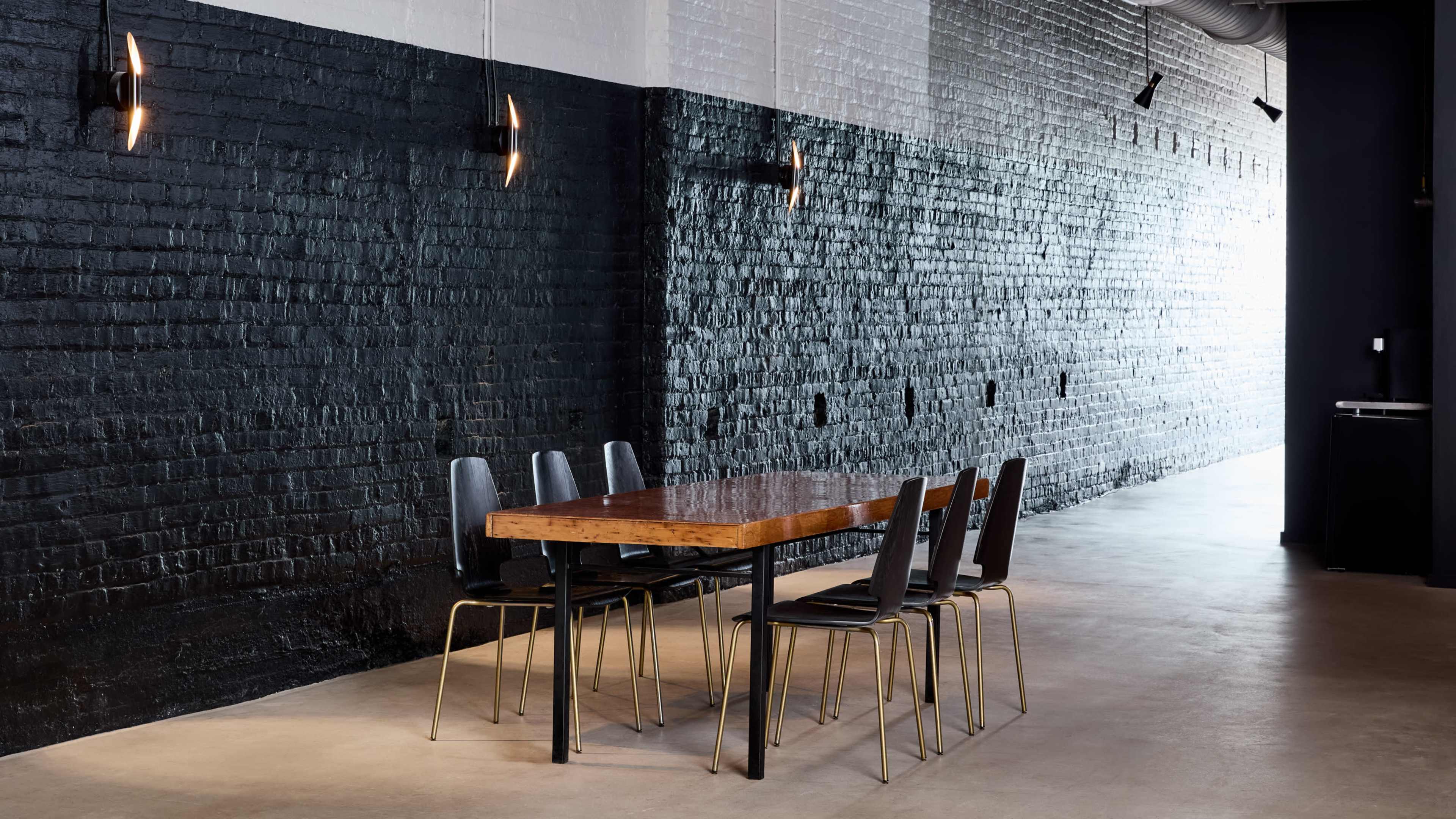 A long wooden table with metal legs is surrounded by several black chairs against a black and white brick wall in a sparsely decorated room.