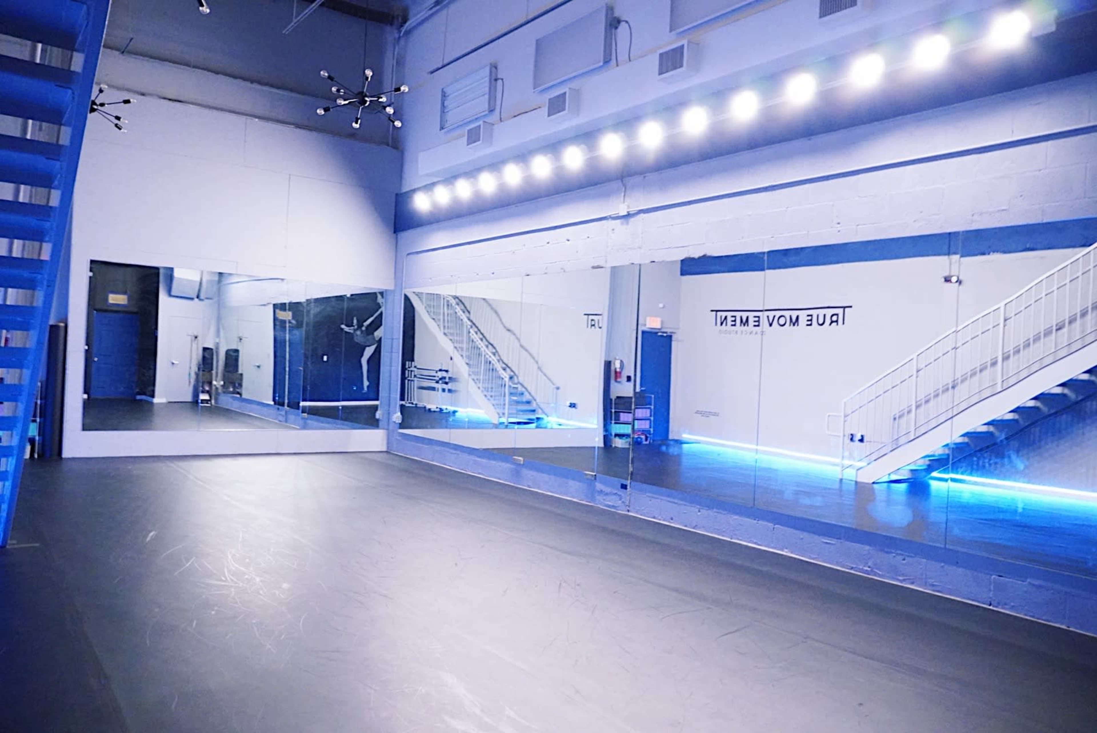 The image shows a spacious dance studio featuring large mirrors, a dark floor, a staircase, and blue LED lighting along the walls.