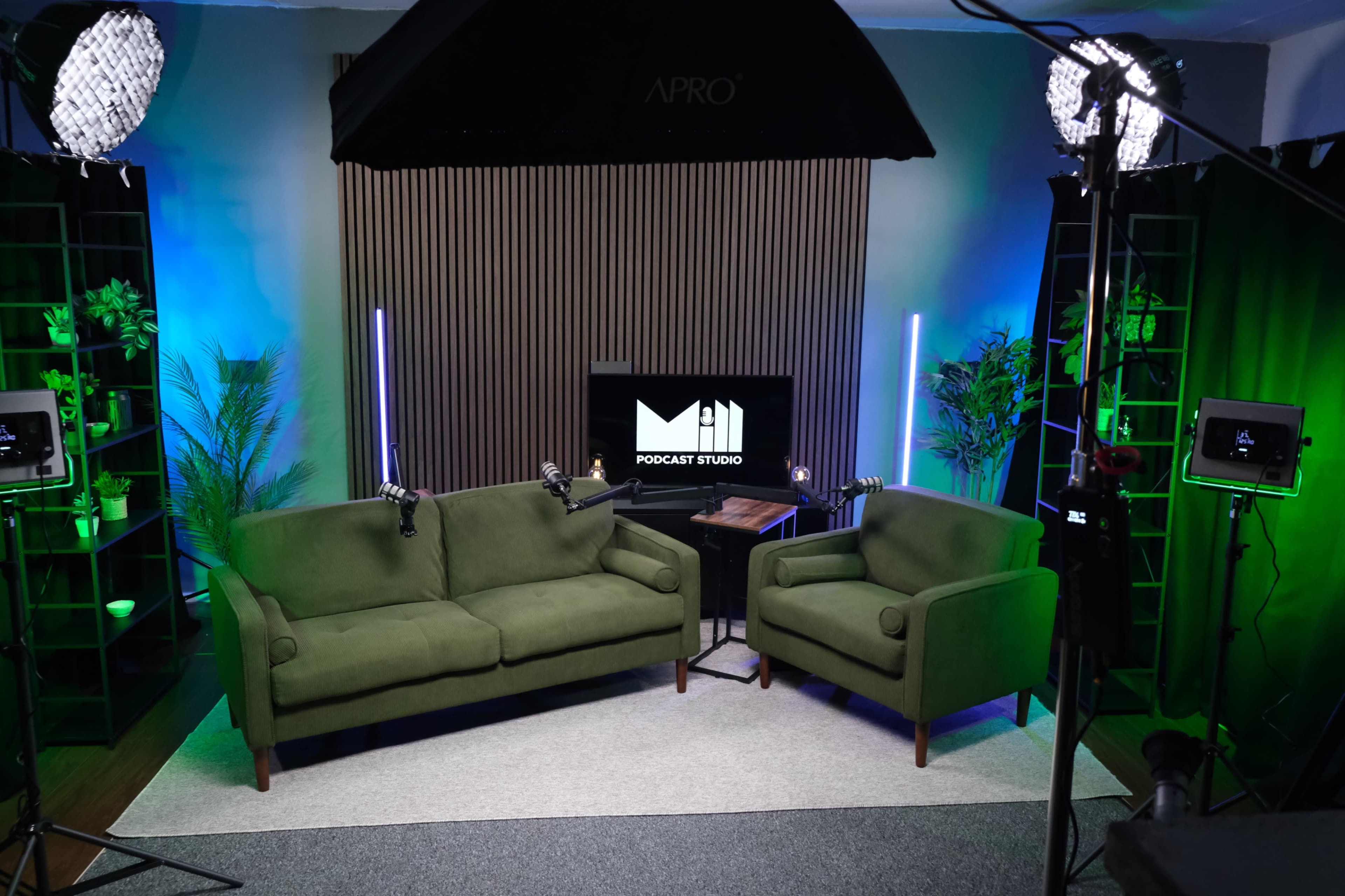 The image shows a podcast studio setup featuring two green sofas, a television screen displaying the words "PODCAST STUDIO," and several lighting equipment arranged around the space.