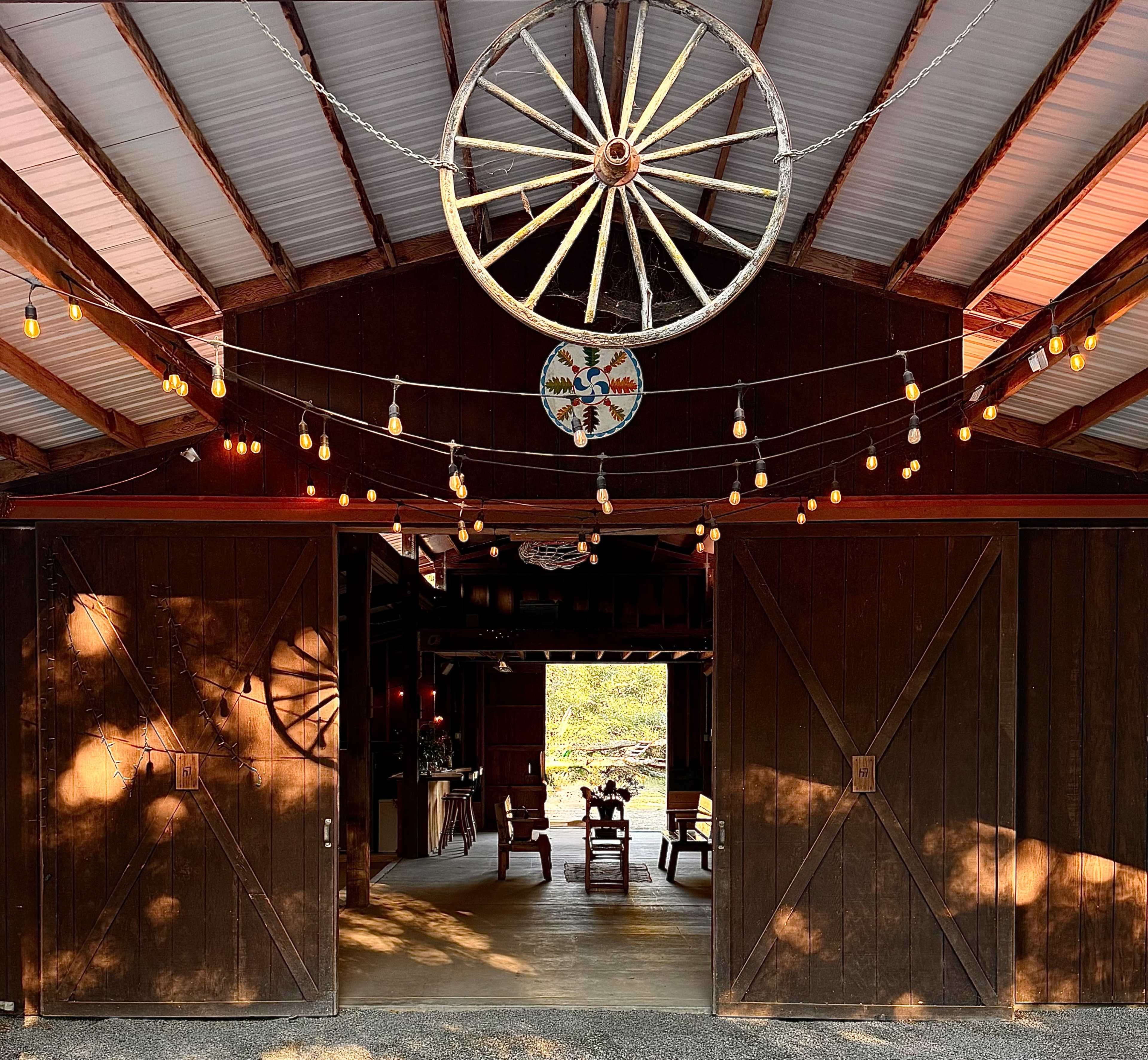 A large wooden barn entrance features a decorative wagon wheel and string lights, leading into a cozy interior with wooden furniture.