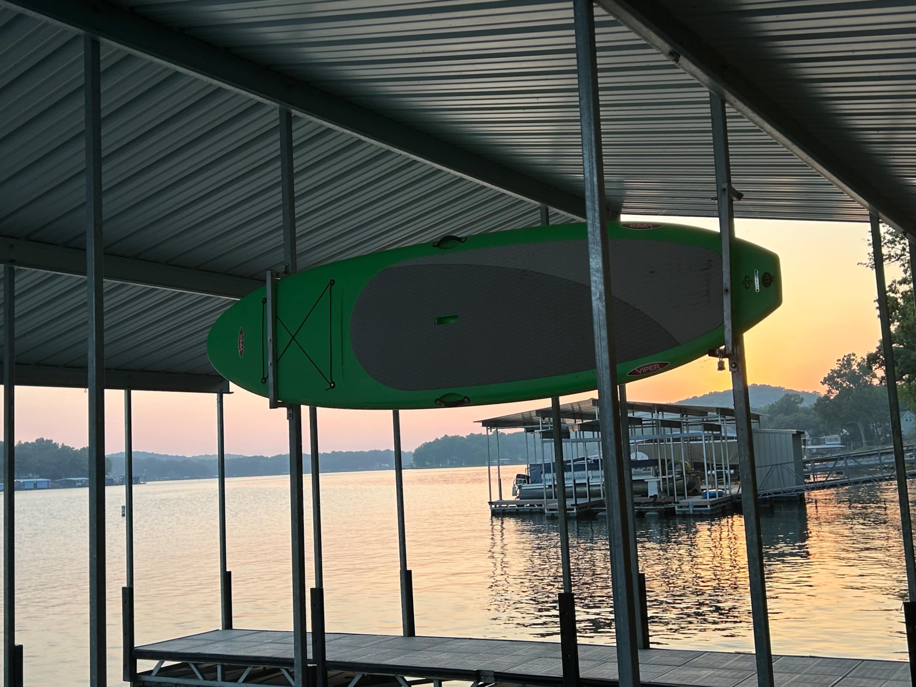 A green paddleboard is suspended from a metal frame under a dock, with a calm lake and a sunset in the background.
