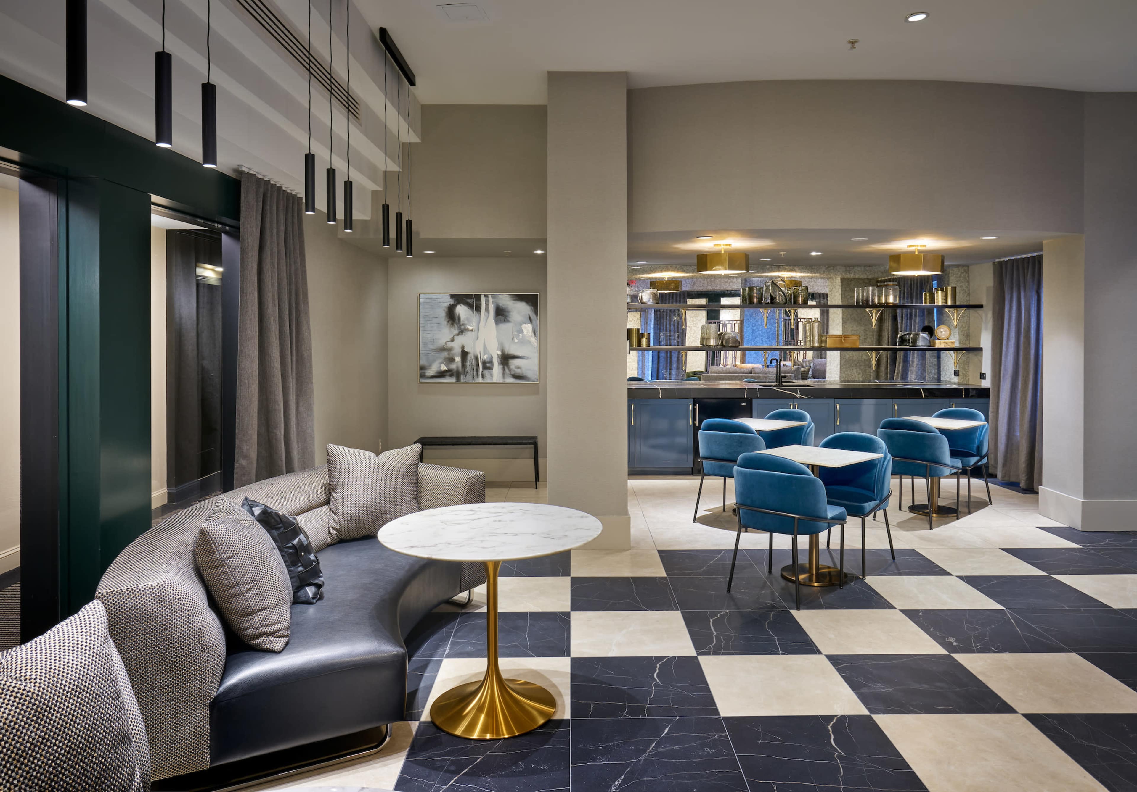 A modern lounge area featuring a curved black sofa, a round marble table, and blue chairs surrounding a nearby dining table, all set against a backdrop of textured walls and ambient lighting.
