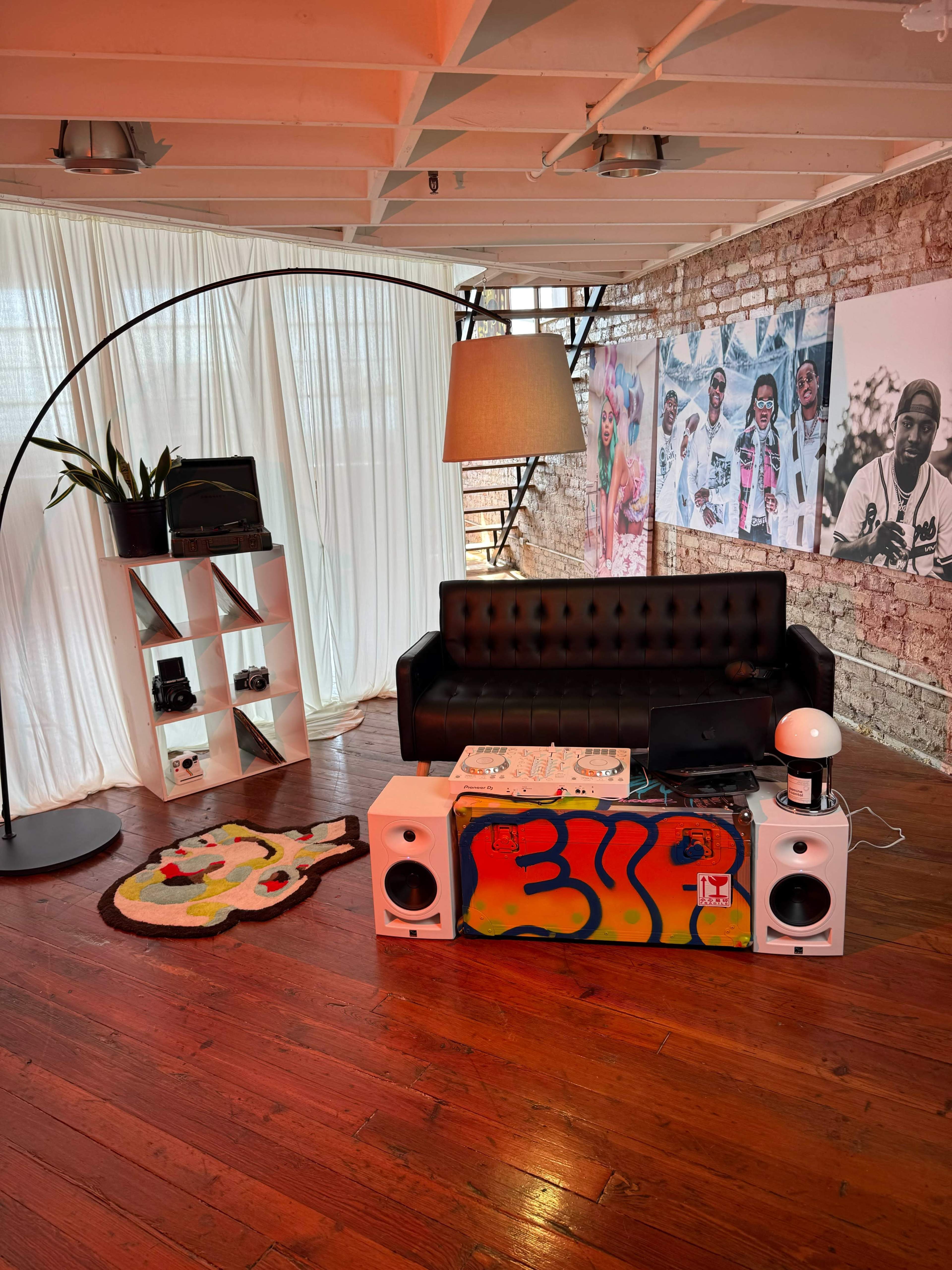 The image shows a modern living space featuring a black leather couch, a uniquely designed coffee table with vibrant artwork, and a tall arc lamp, all against a backdrop of exposed brick walls and large photographs.