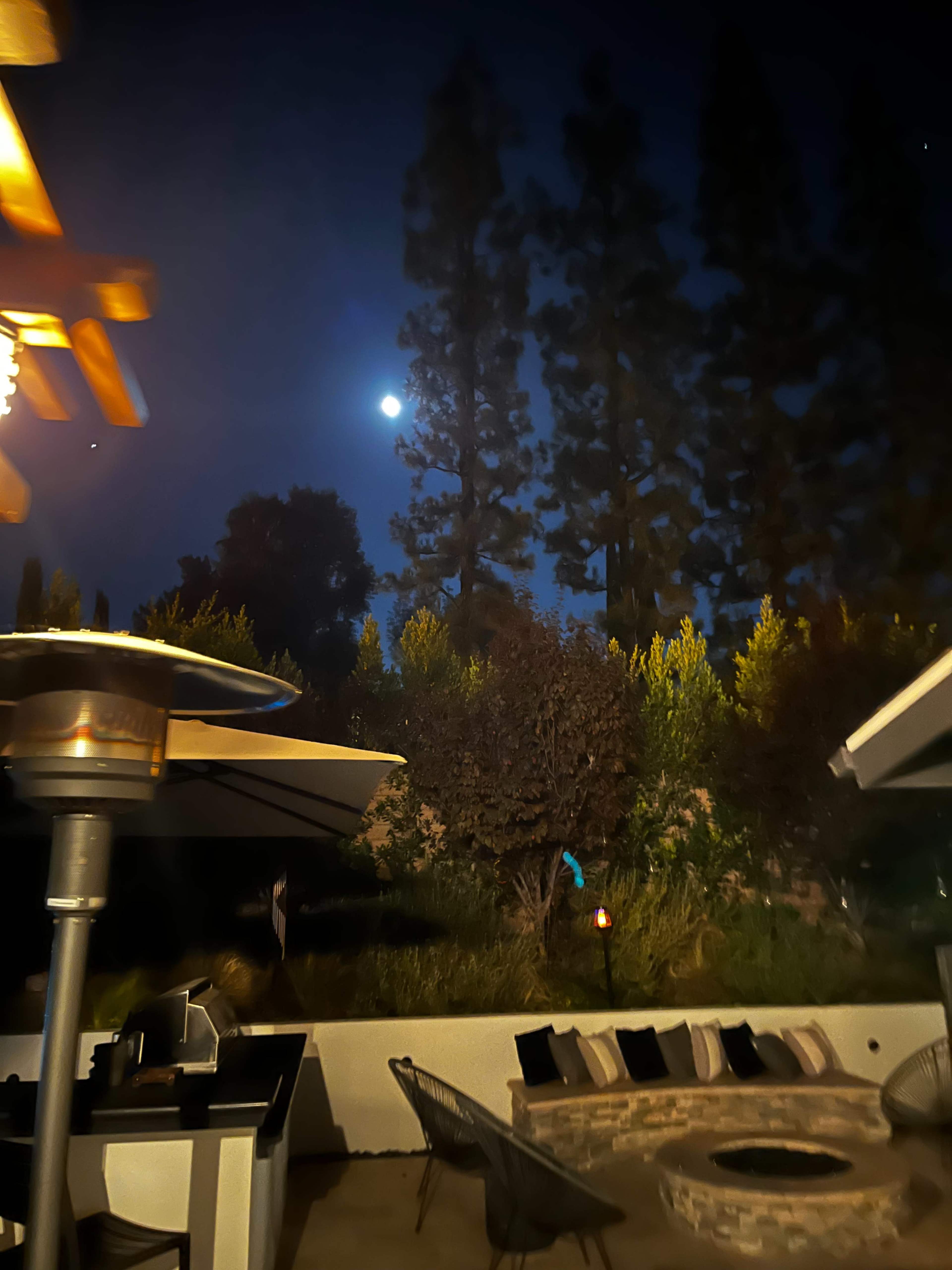 A bright full moon illuminates a backyard surrounded by tall trees, with outdoor seating and a fire pit visible.