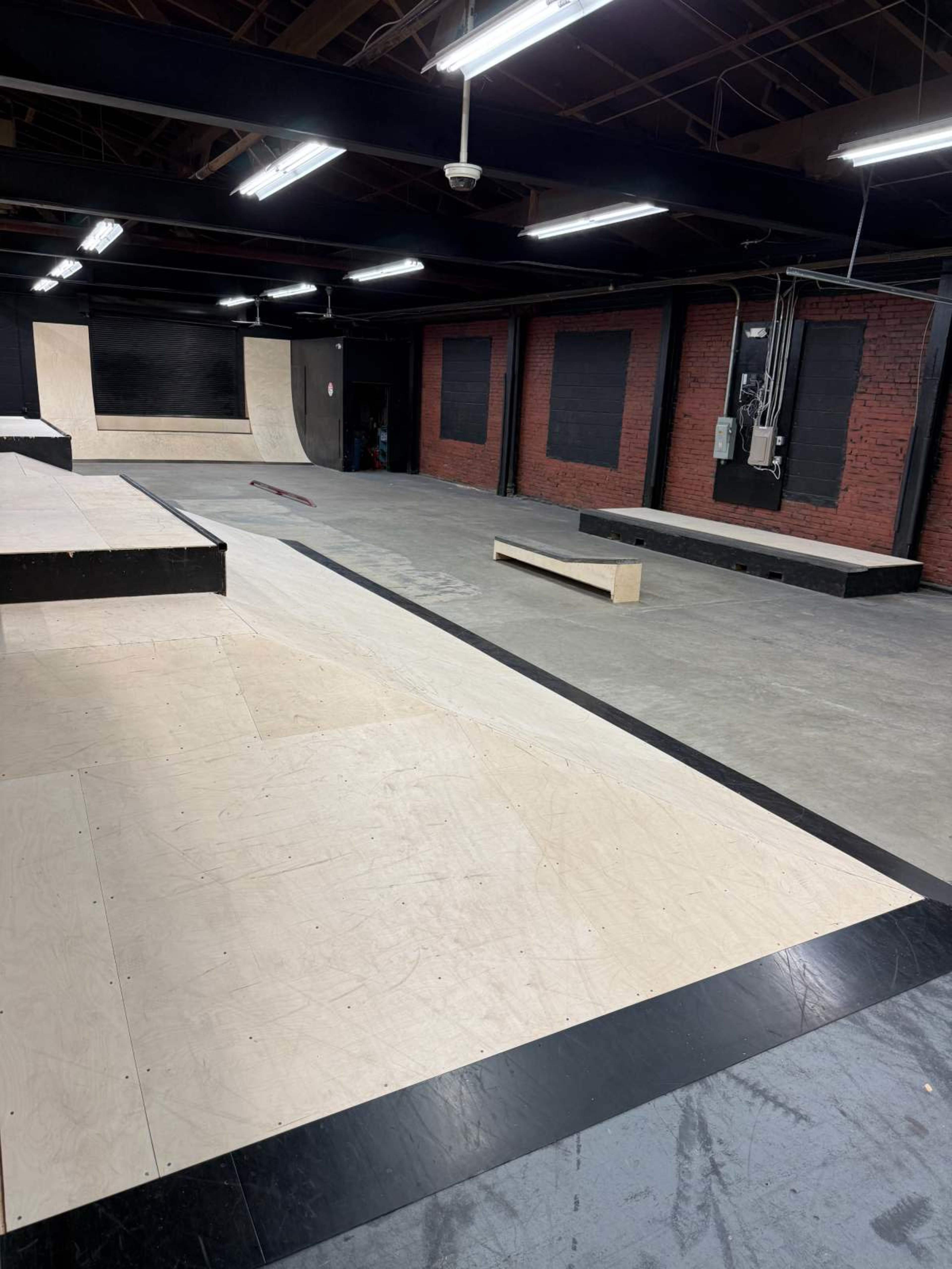 The image shows an interior skate park with ramps, flat surfaces, and a wooden box for tricks under bright overhead lights.