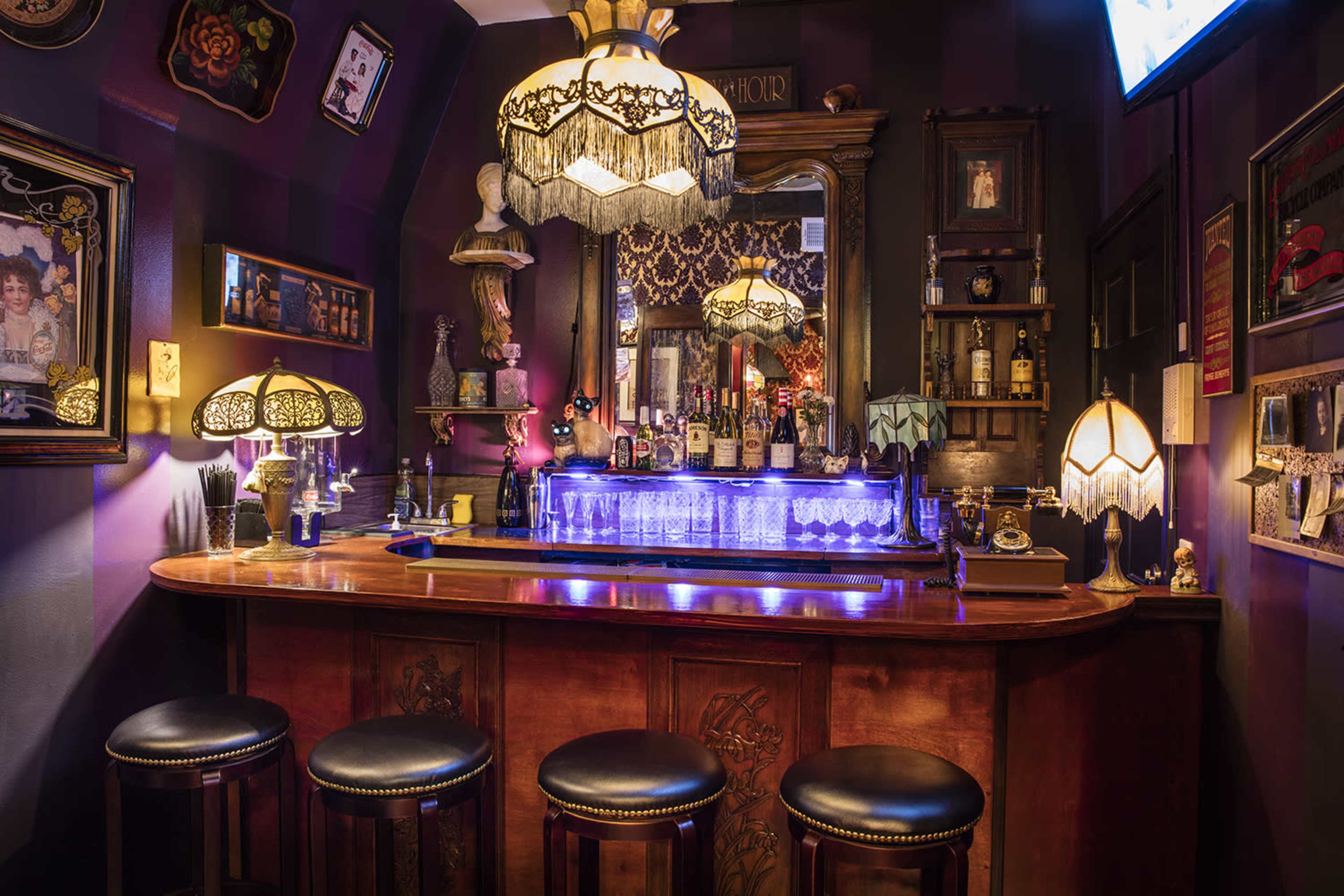 A dimly lit bar featuring a wooden counter with four black leather stools and a mirrored back wall adorned with ornate decor and illuminated by vintage-style lamps.