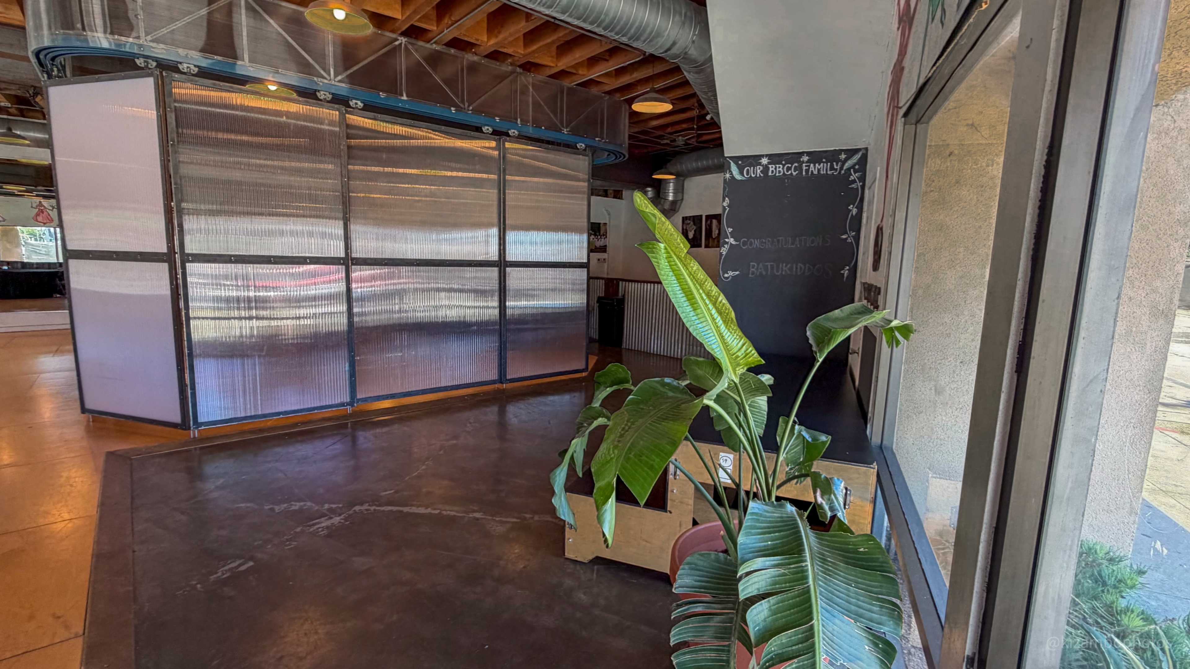 The image shows an interior space featuring a plant, a glass partition, and a chalkboard against a backdrop of wooden beams and polished concrete flooring.